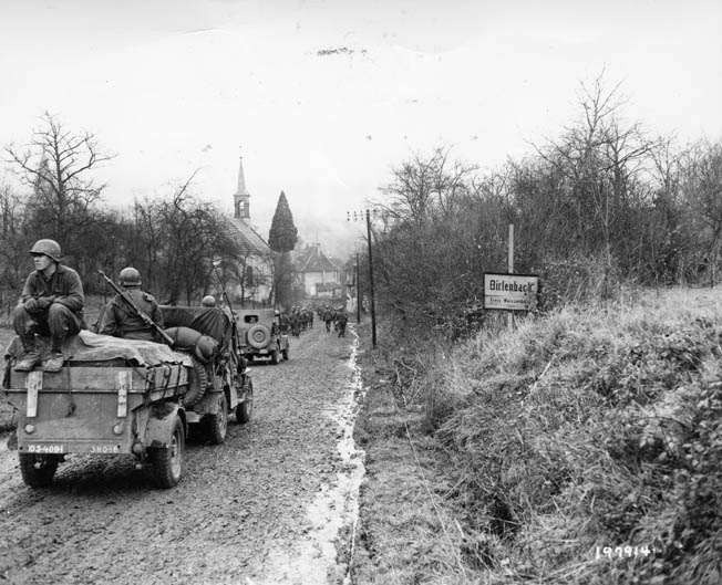 On December 14, 1944, two days before the opening of the Battle of the Bulge, infantrymen of the 103rd Division ride atop jeeps and trailers as they move into the town of Birlenbach, France. The 103rd crossed into Germany the following day and began its assault on the Siegfried Line.