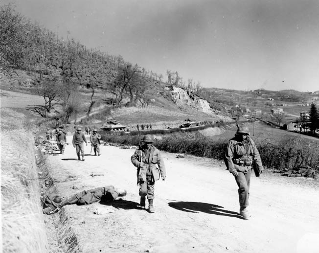 A line of 10th Mountain troops, accompanied by armored vehicles, passes dead Germans along a road in the northern Apennines, March 4, 1945.