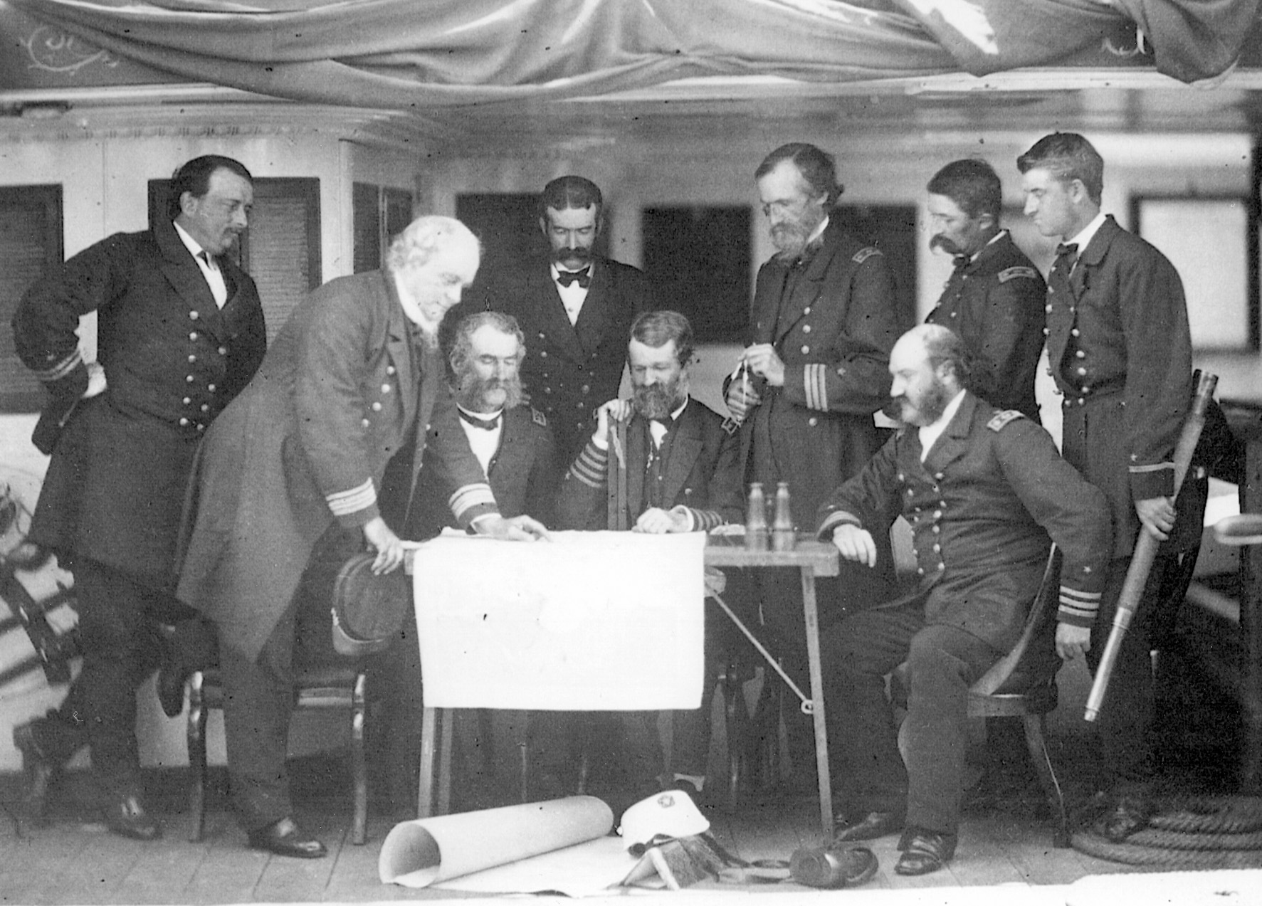 A council of war on the USS Colorado. Rear Admiral John Rogers has one hand on the chart, another on his cap.