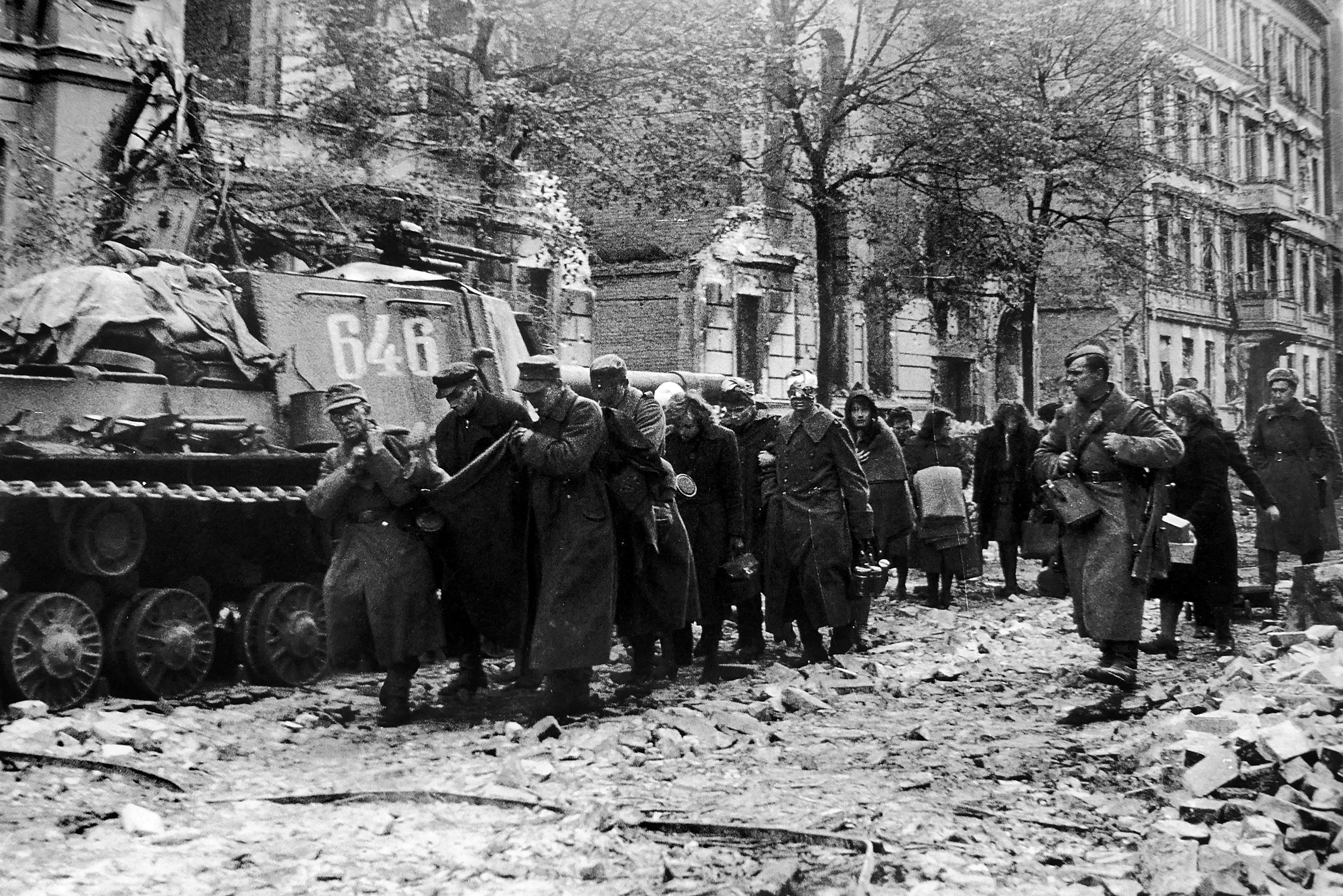 German prisoners carry a wounded comrade in a blanket as they make their way down a Berlin street in the spring of 1945. These captives are walking past a Soviet ISU-152 self-propelled gun.