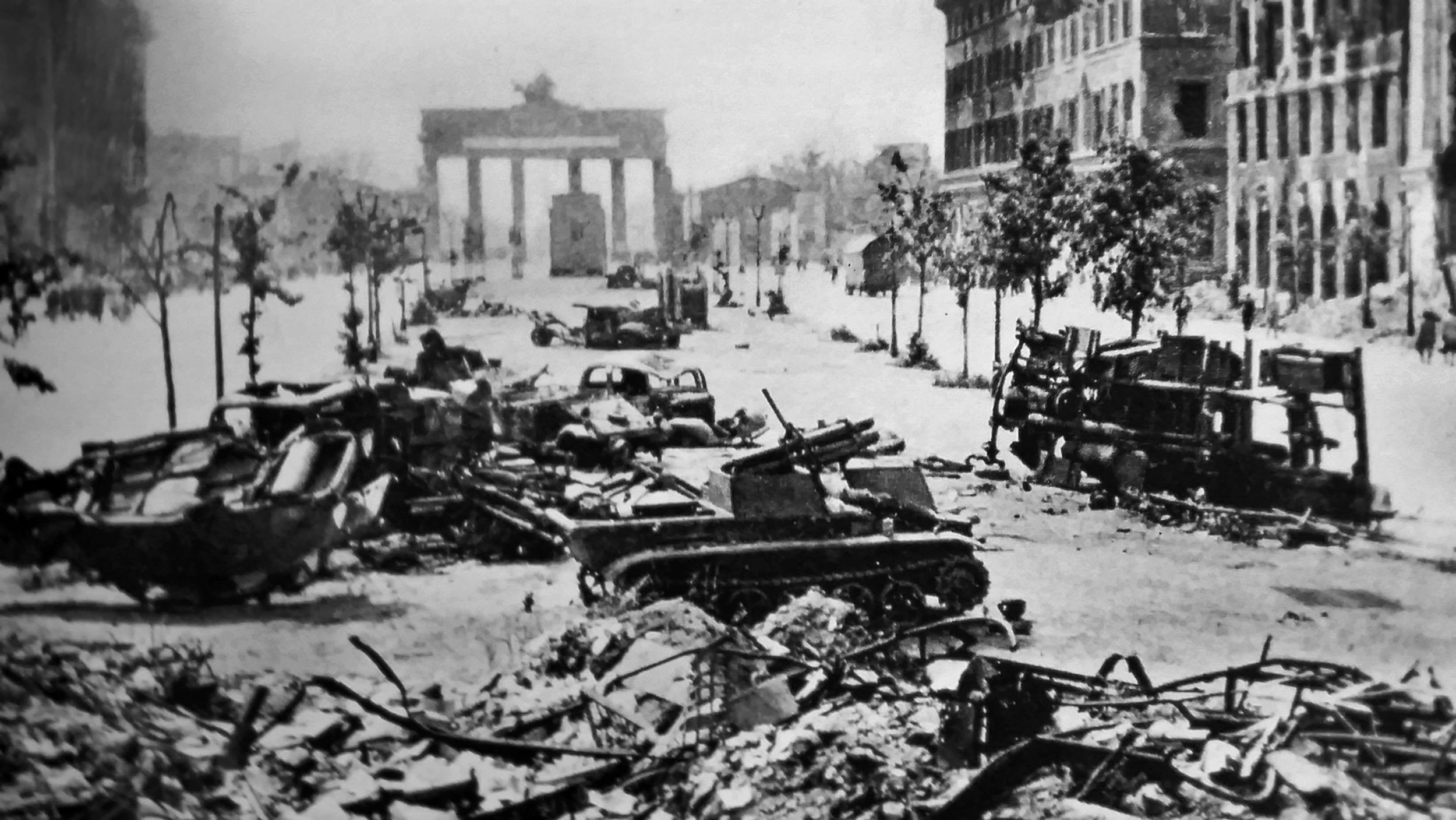Demolished vehicles and damaged buildings are shown in this stark landscape of Berlin shortly after the battle in the spring of 1945. The Brandenburg Gate looms in the background.