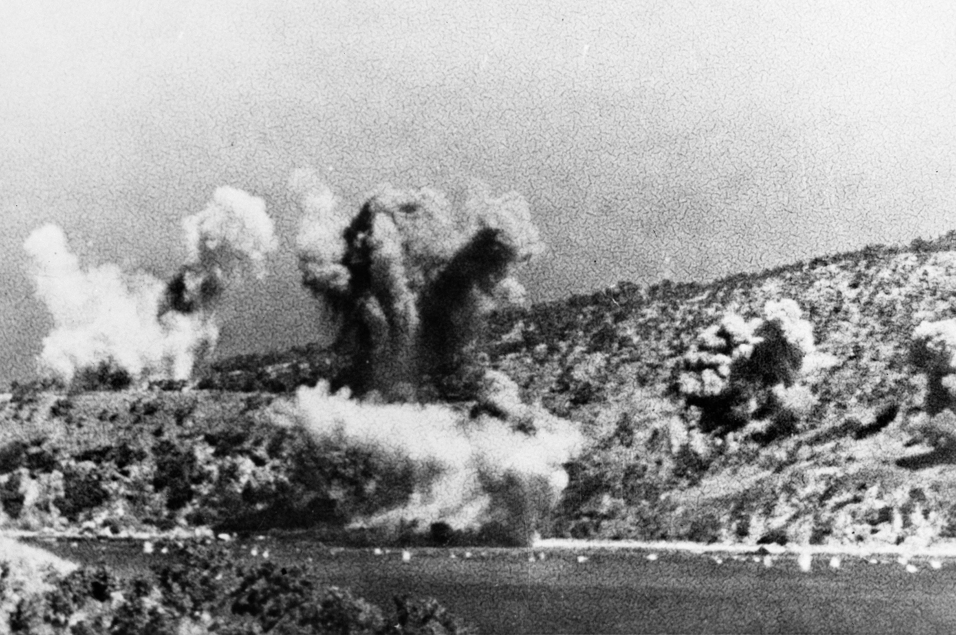 Pillars of smoke rise from the hills and harbor as Japanese aircraft carry out an air raid on Allied shipping targets in Milne Bay.