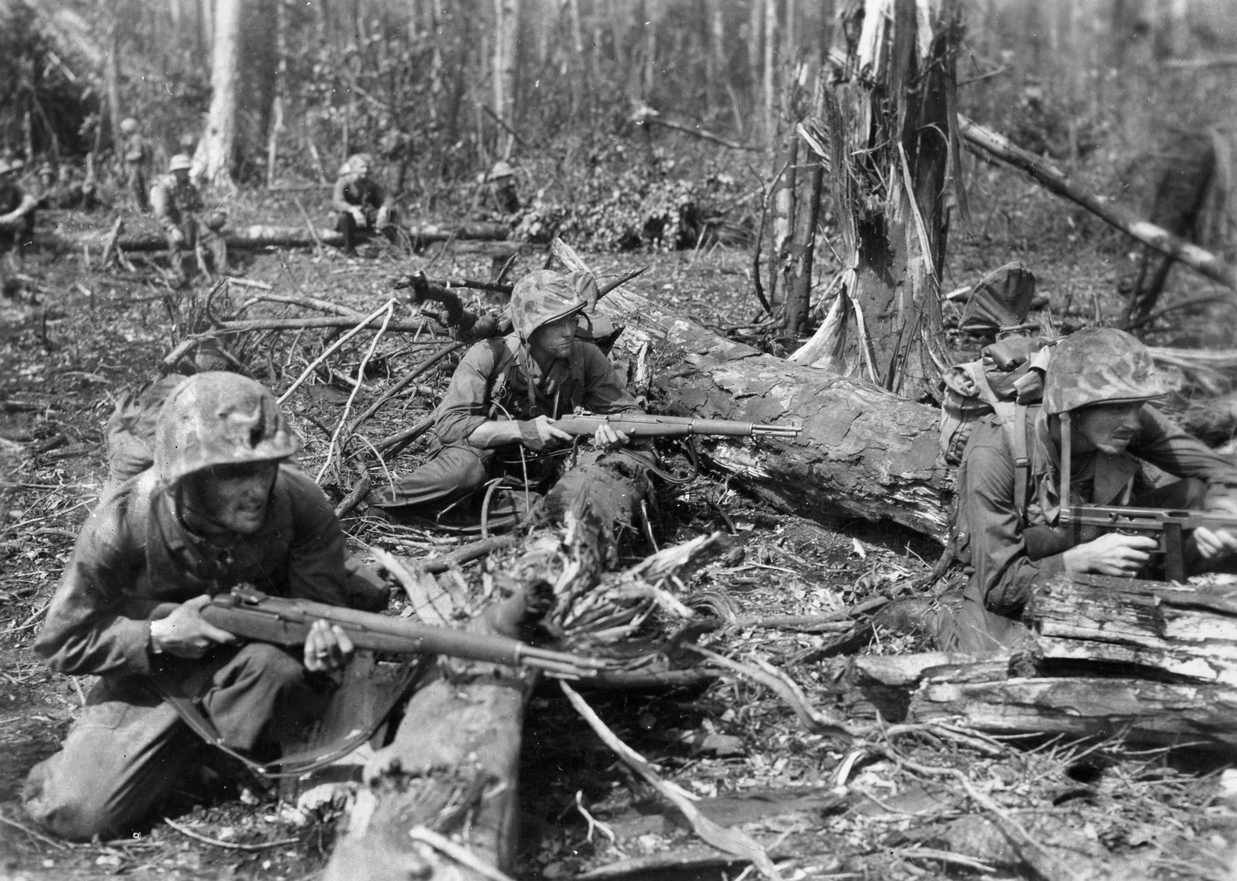 During action on the first day of fighting at Cape Gloucester, three U.S. Marines take cover from Japanese fire among fallen palm trees. Two of the Marines are armed with M-1 rifles, while the man on the right carries a Thompson submachine gun.