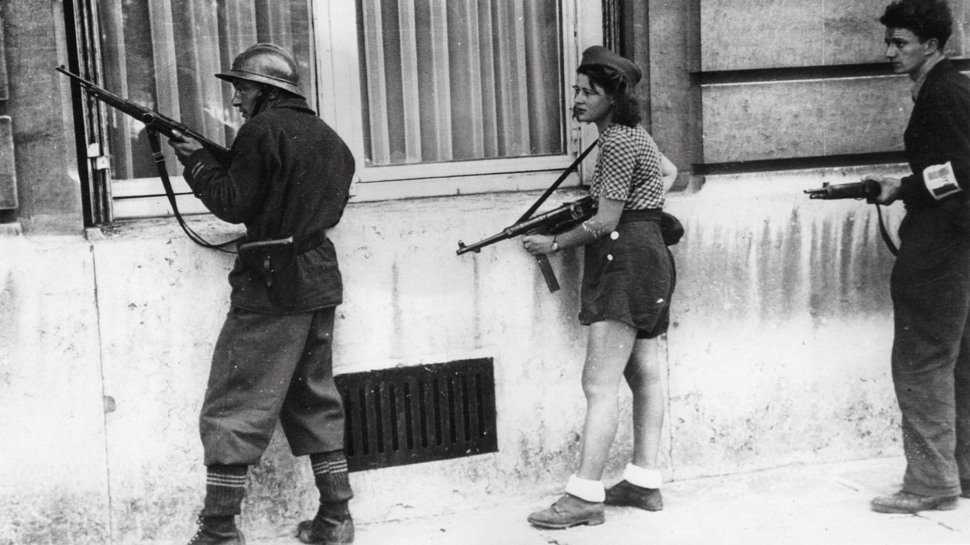 A young French woman carries a captured German weapon and moves with a comrade alongside a building in Paris.