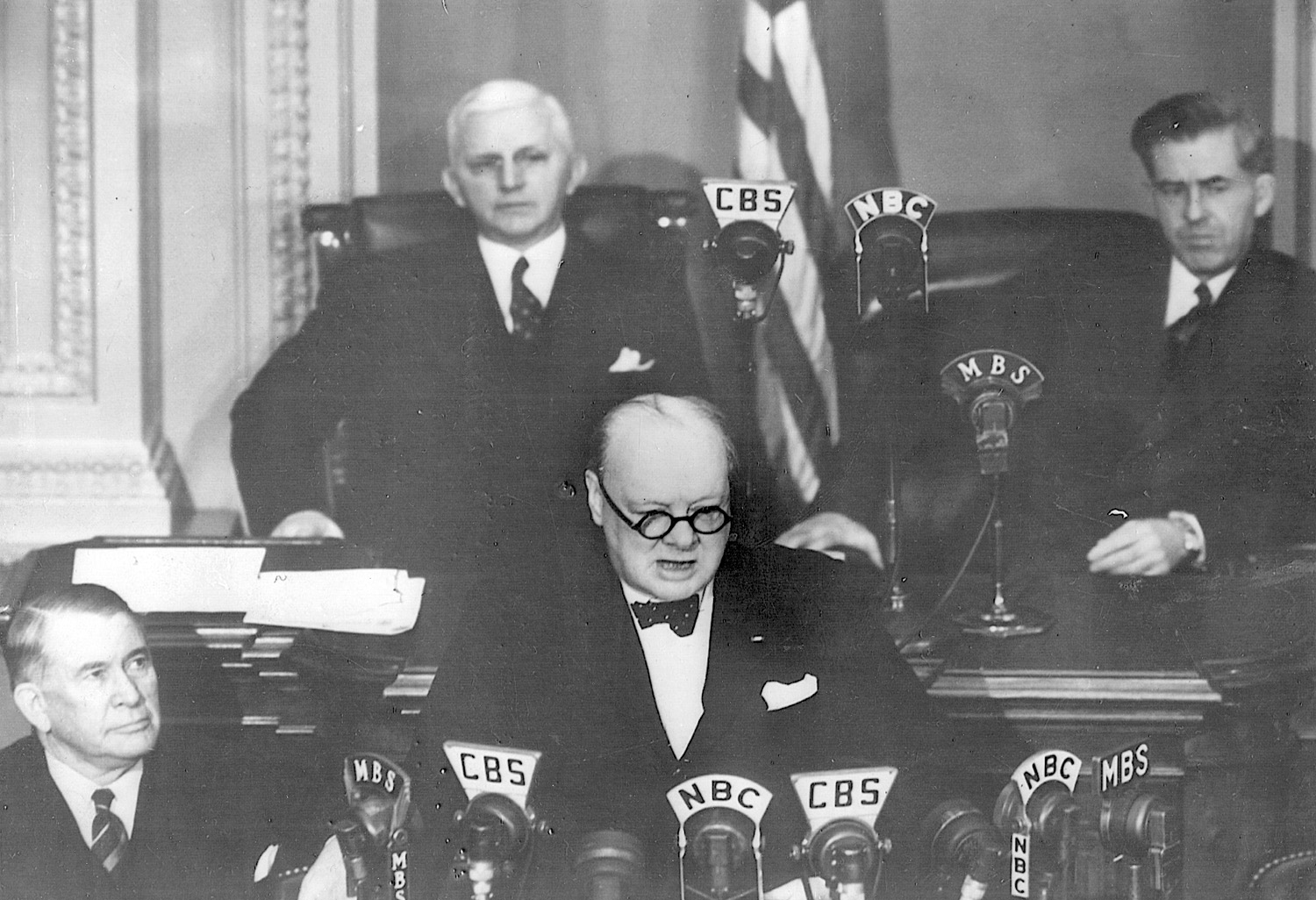 British Prime Minister Winston Churchill addresses a joint session of Congress on December 26, 1941. Behind him are Representative William P. Cole, Jr., (D-Md.), House speaker pro tem (left), and Henry A. Wallace (right). Below sits Senate Majority Leader Alben W. Barkley (D-Ky.). 