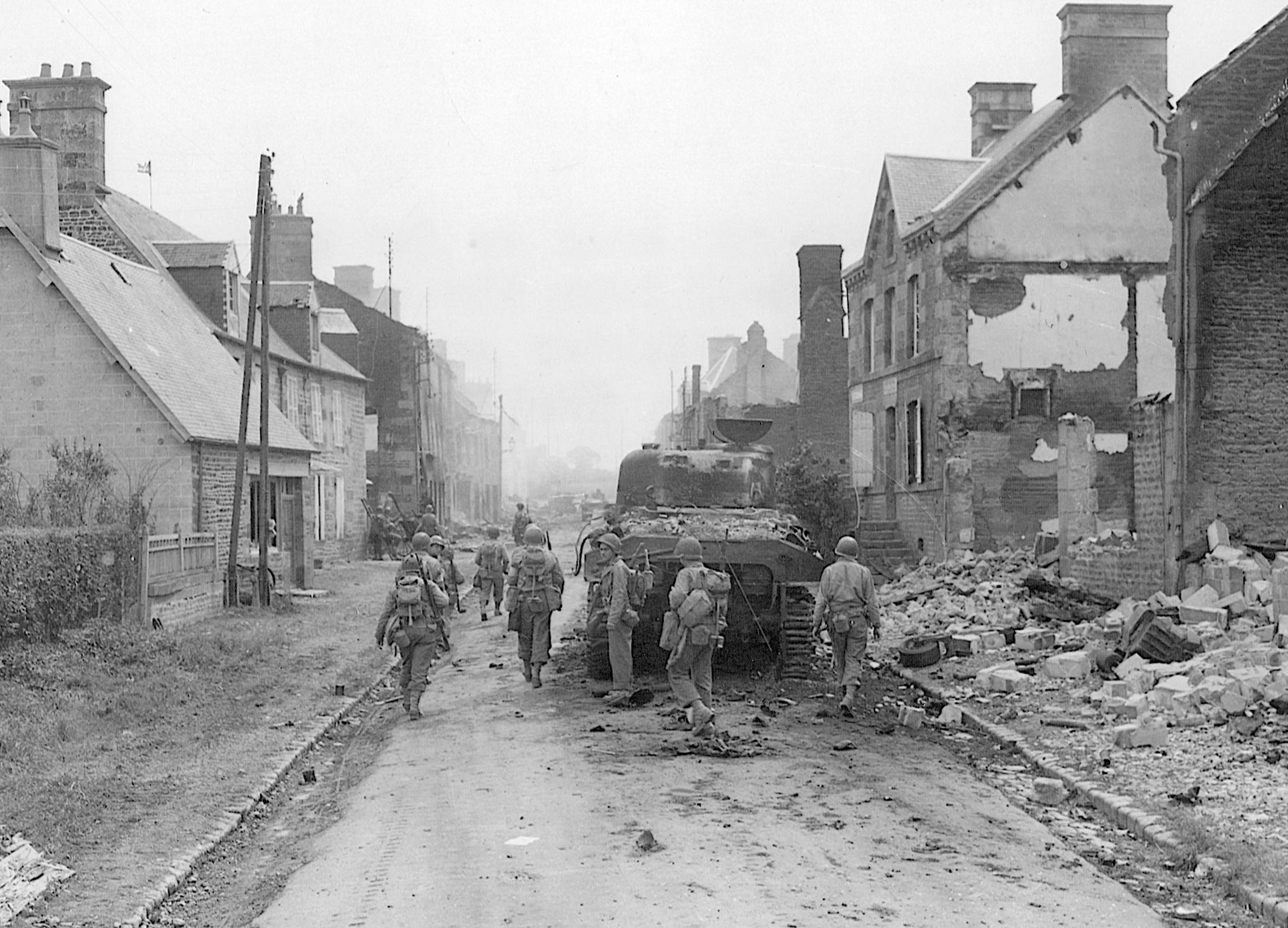 GIs trudge past the bulk of a Sherman tank knocked out during street fighting on August 3, 1944, as they advance toward the fighting at the strategic town of Mortain. The 30th Division lost approximately 1,800 men killed and wounded at Mortain, 277 of these defending Hill 314.