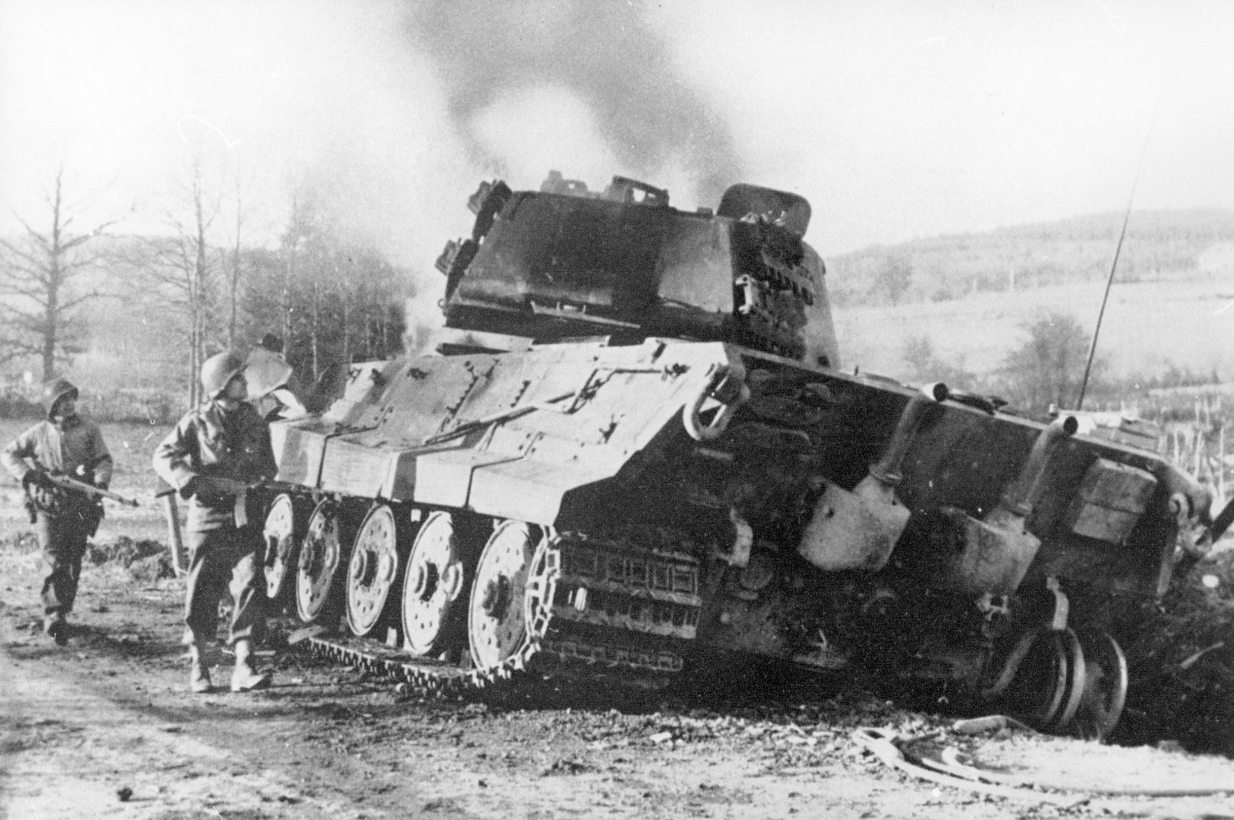 The hulk of a German Tiger tank, destroyed December 18, 1944, still smolders as a pair of American GIs ambles warily past, east of La Gleize. 