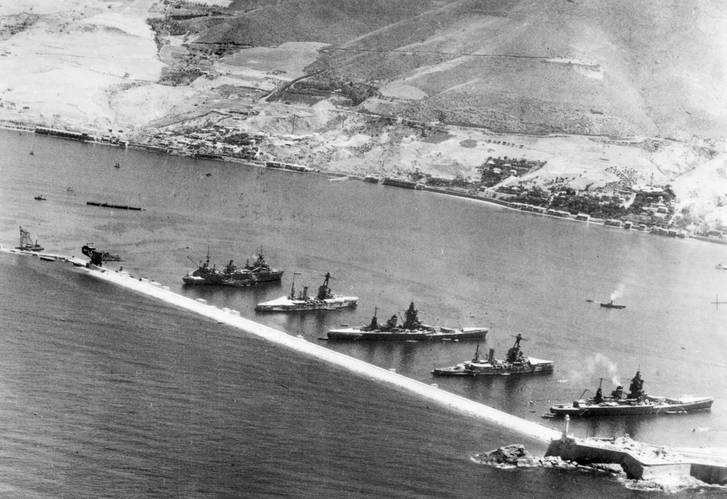 Five powerful French warships align stern-first to the mole at Mers-el-Kébir before the Royal Navy attack. Lesser ships align to the right. 