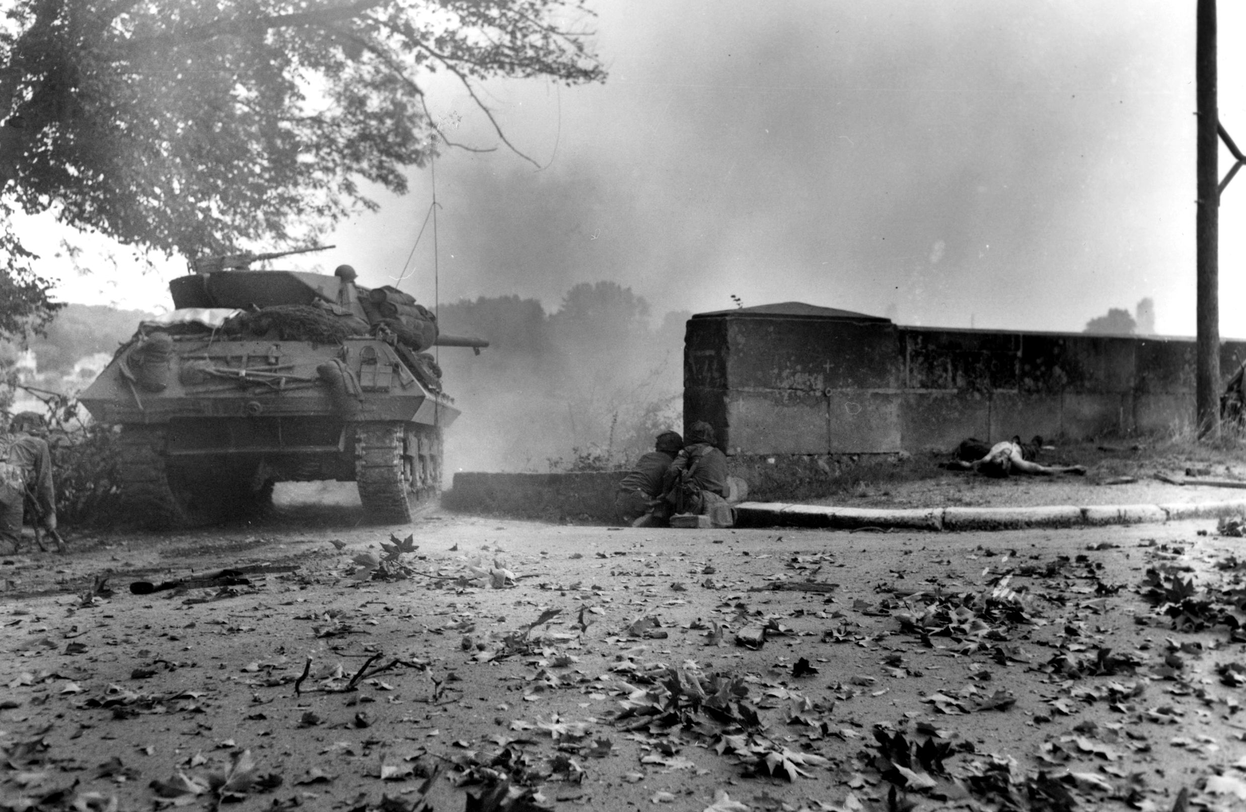 An American tank destroyer tentatively approaches the smoldering ruins of a Normandy town as accompanying infantrymen take cover from incoming German fire.