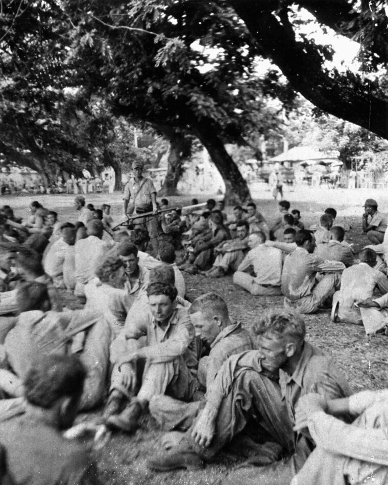 During the arduous march from the Bataan Peninsula to Cabanatuan prison, American prisoners are allowed a brief respite from the dust and heat. A Japanese soldier, with fixed bayonet, watches the captives, many of whom would die along the way. 