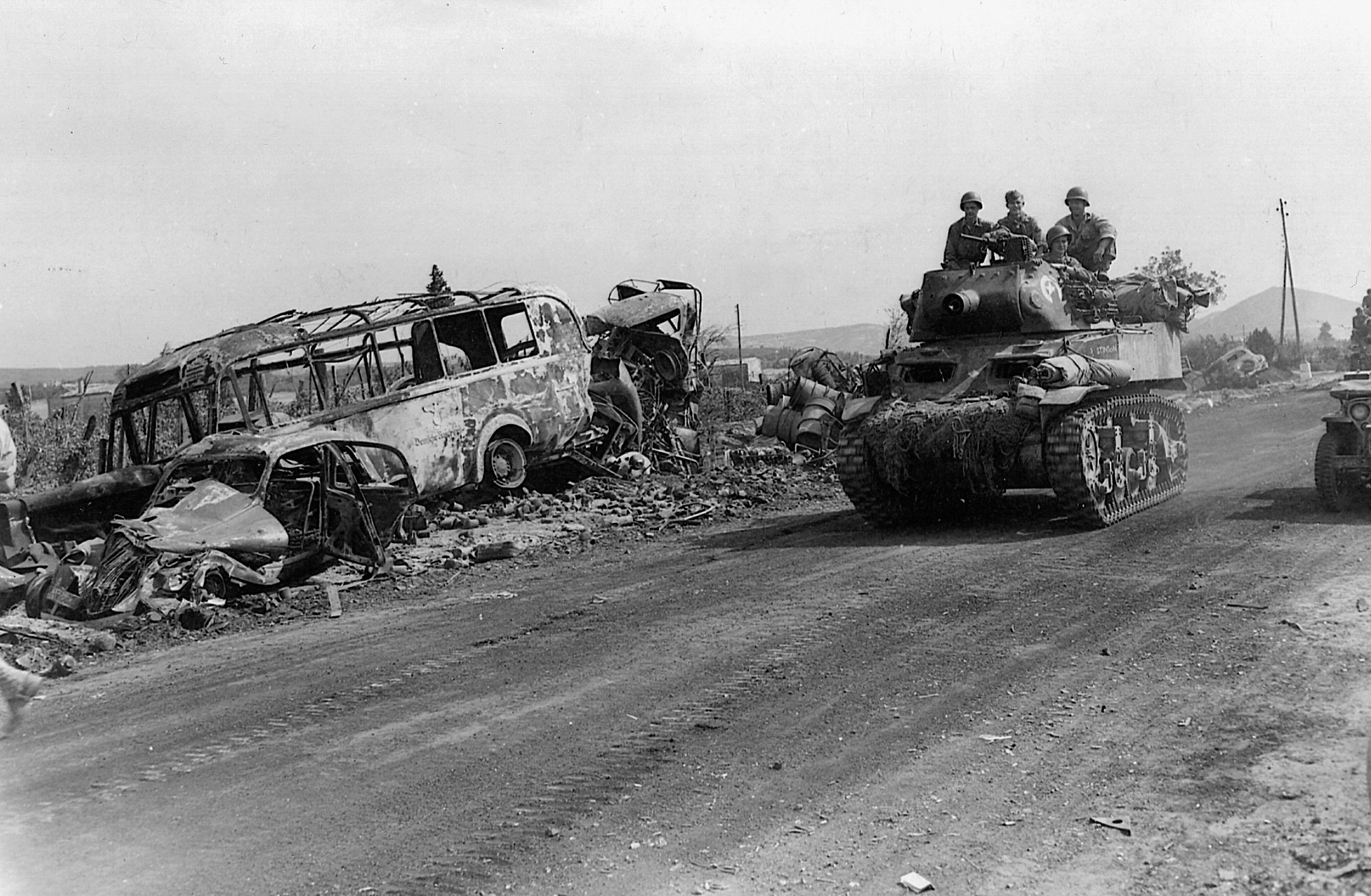 Passing the charred remnants of a bus, tanks of the U.S. 7th Army roll down a dusty road into the town of Montelimar.