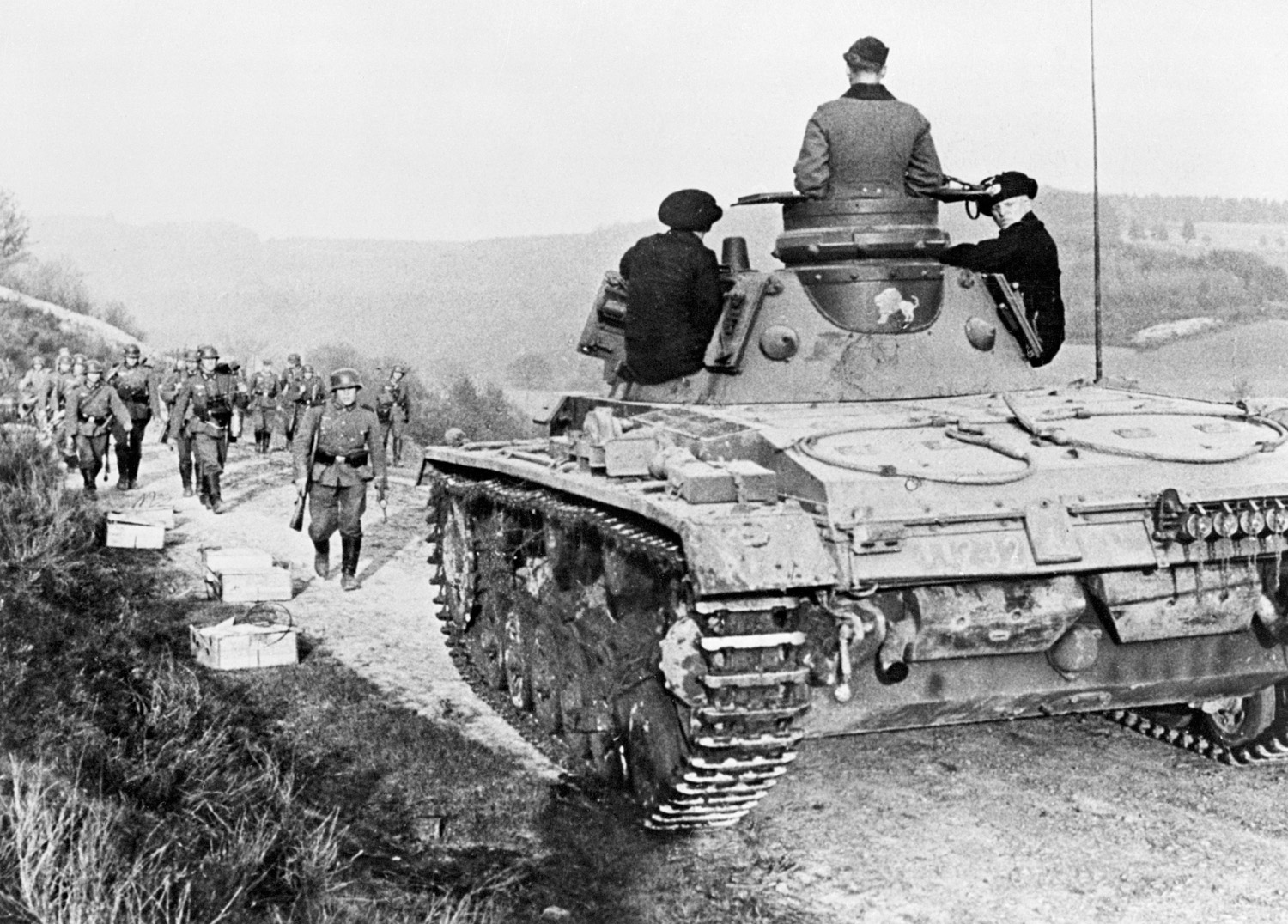 With a steady stream of infantry following behind, a German tank makes its way up a dusty French road.