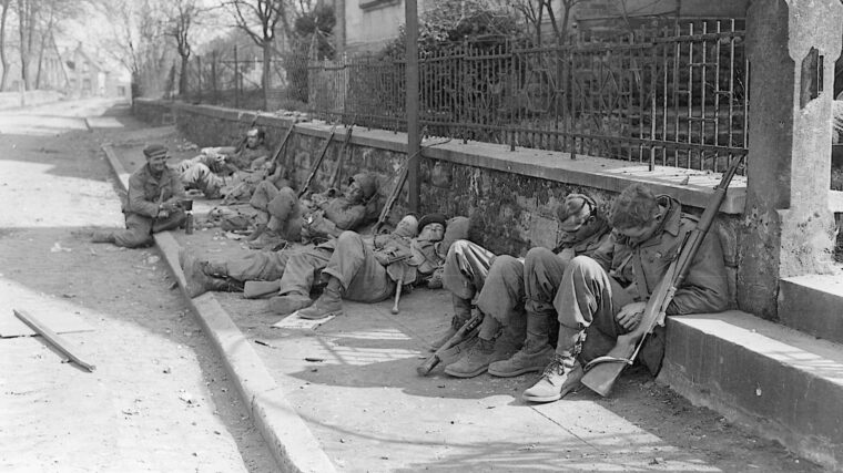 After fighting all night and clearing over 100 pillboxes in the Siegfried Line, men of the 36th Infantry Division collapse on a sidewalk in Berg Zabern, Germany. The end of the war is a month away.