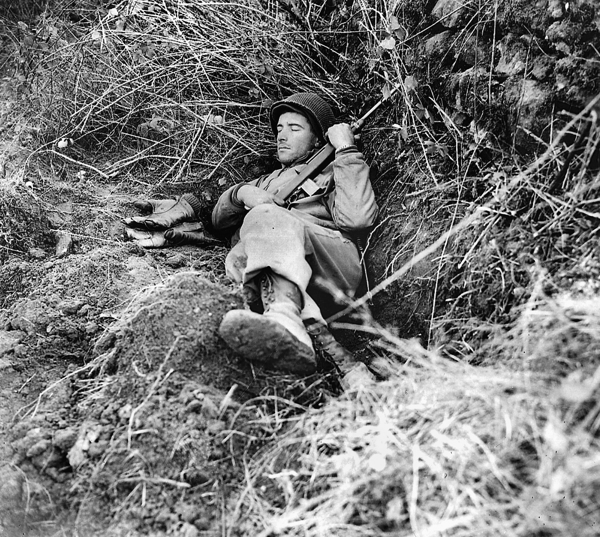 An infantryman, carbine still in his grip, snoozes in a slit trench while waiting to move to the front. 