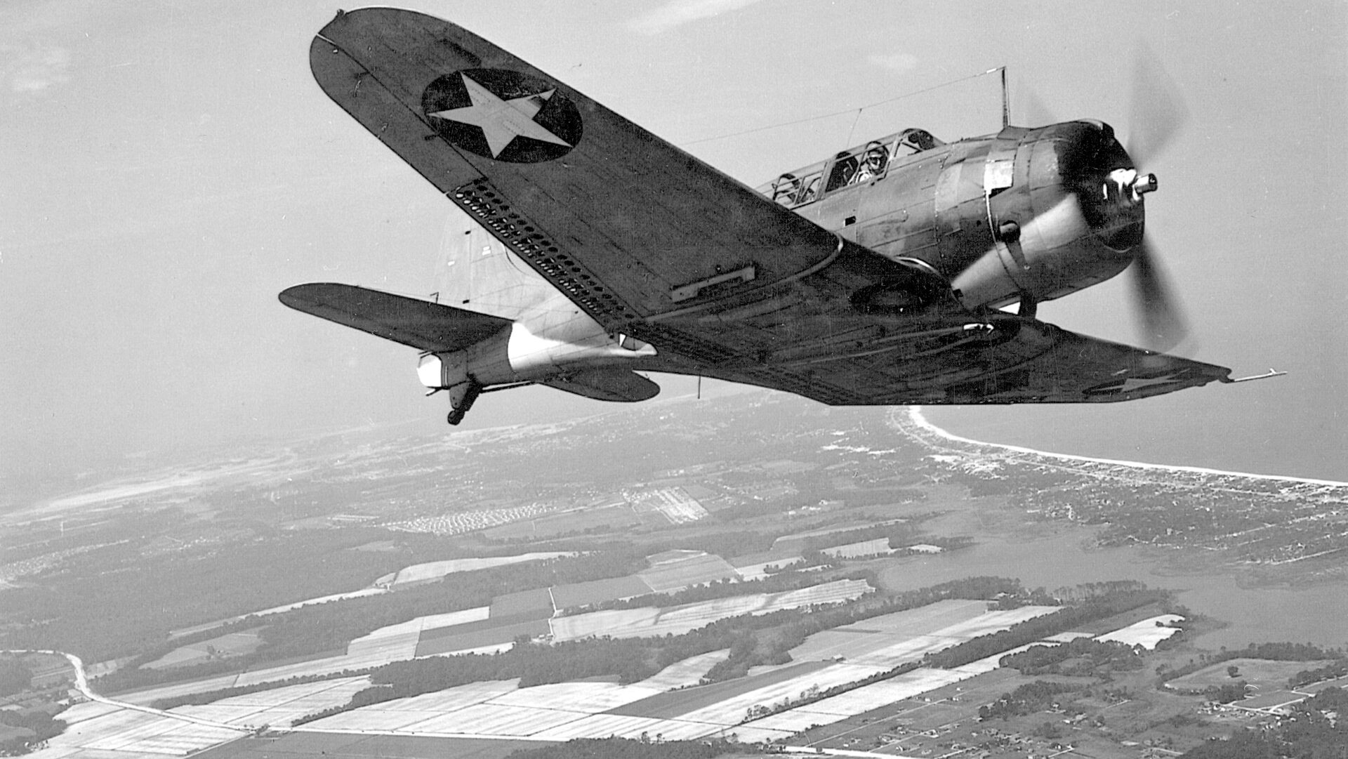 An SBD flies over the environs of Norfolk, Va. Visible are the split dive brakes at the trailing edge of the wing so important to the stability of the aircraft in a dive-bombing run on an enemy ship.