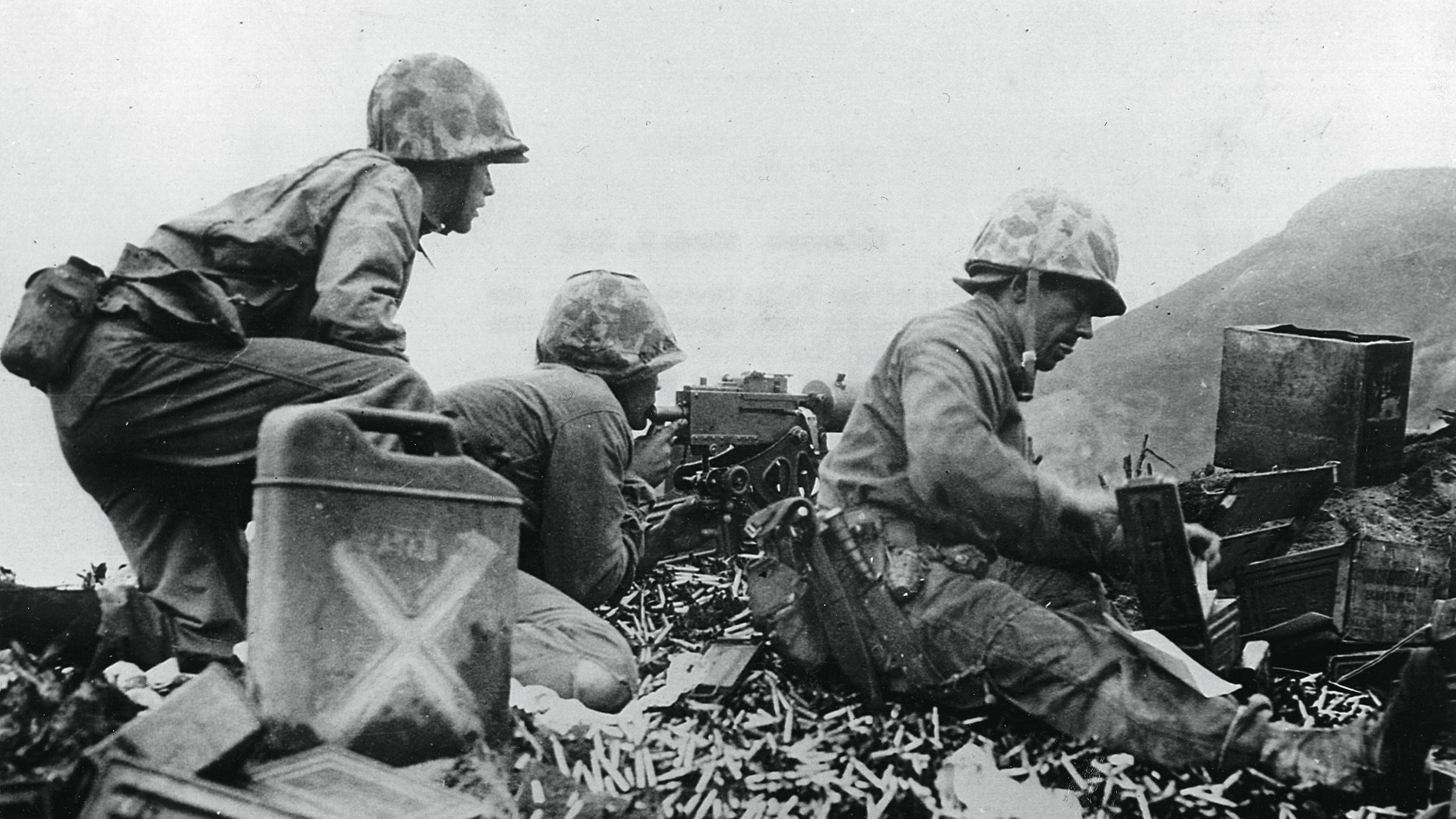 With their Ka-Bar fighting knives at their sides, U.S. Marines sit atop a pile of spent shells and provide cover for comrades moving inland on Iwo Jima.