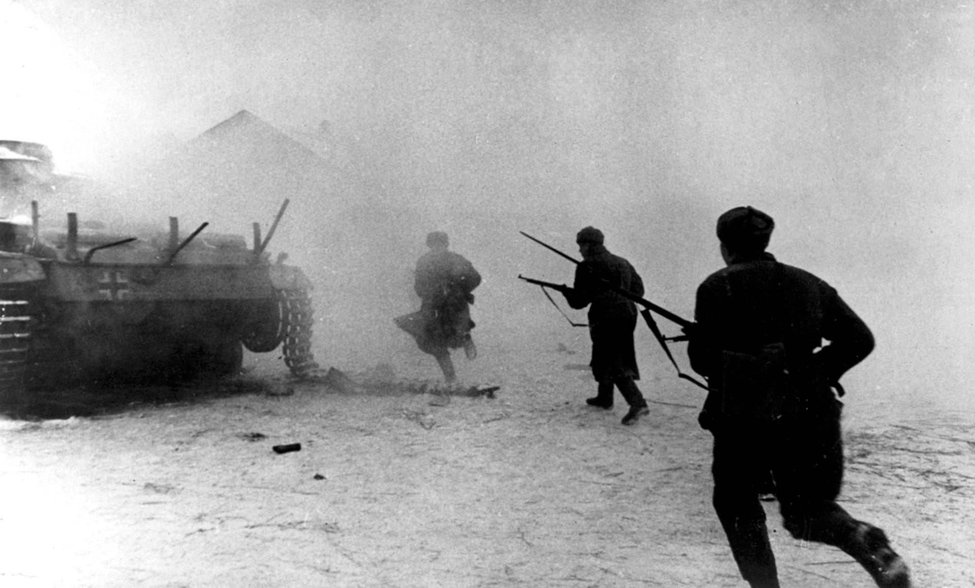 Hurrying past a smoldering German tank, Red Army soldiers plunge into the haze of combat with their rifles at the ready.