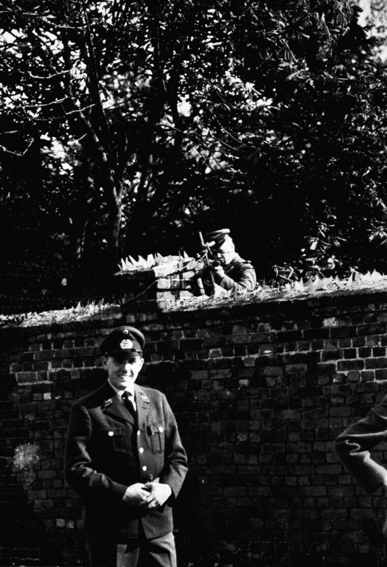 A West Berliner poses in uniform while an East German guard aims his gun menacingly over the wall.