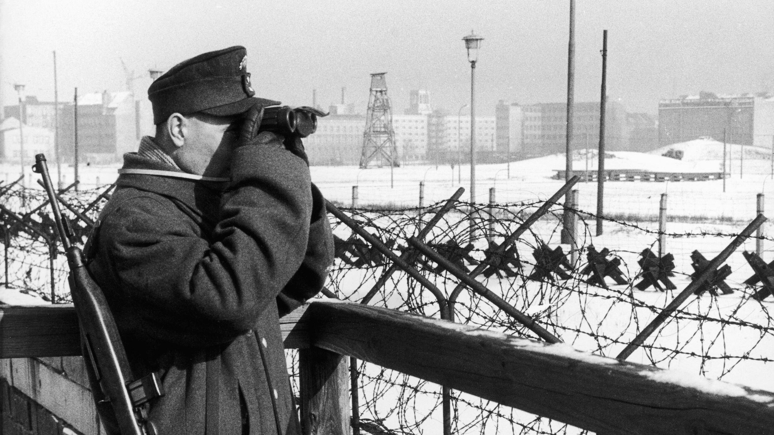 An armed West German policeman keeps a close watch on the East Zone across the Berlin Wall. The nearest mound of snow marks the remains of Adolf Hitler’s underground bunker.