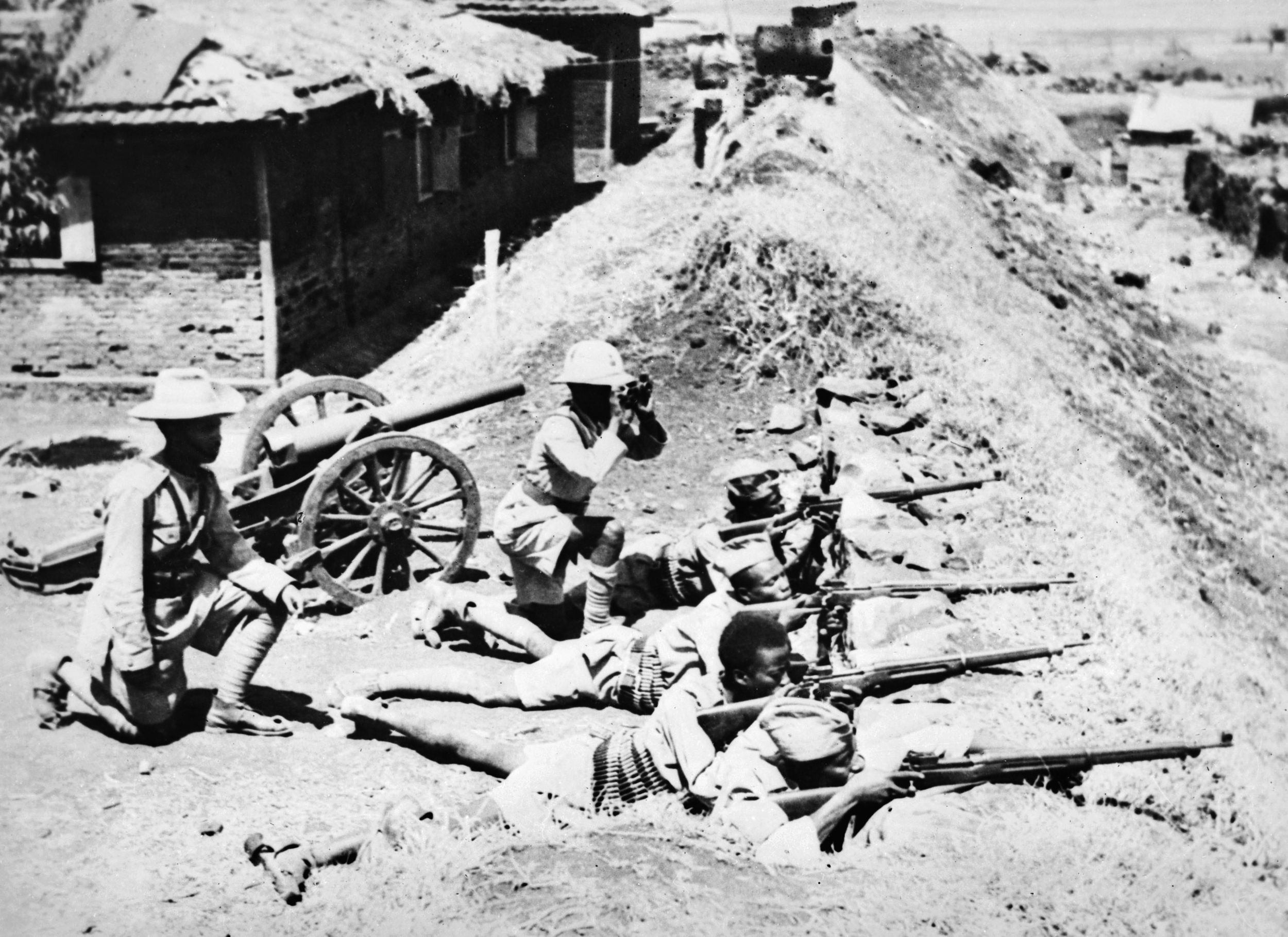 One officer directs riflemen into position, while another scans the horizon through binoculars as Abyssinian soldiers man positions along the rampart of a fort in Ethiopia. An antiquated artillery piece sits nearby.