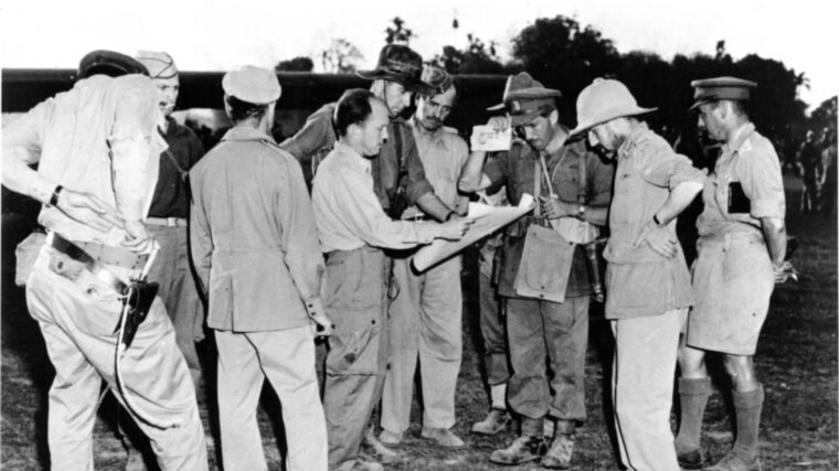 In this classic photograph, Brigadier General Orde Wingate is next to Brigadier Derek Tulloch at the far right. Majors Philip Cochran (back facing camera) and John Allison are on the left. Major Alison is holding a map with Brigadier Scott, who wrote an account of this informal gathering. Air Marshal Baldwin and Brigadier Michael Calvert stand between Scott and Wingate. (Imperial War Museum)