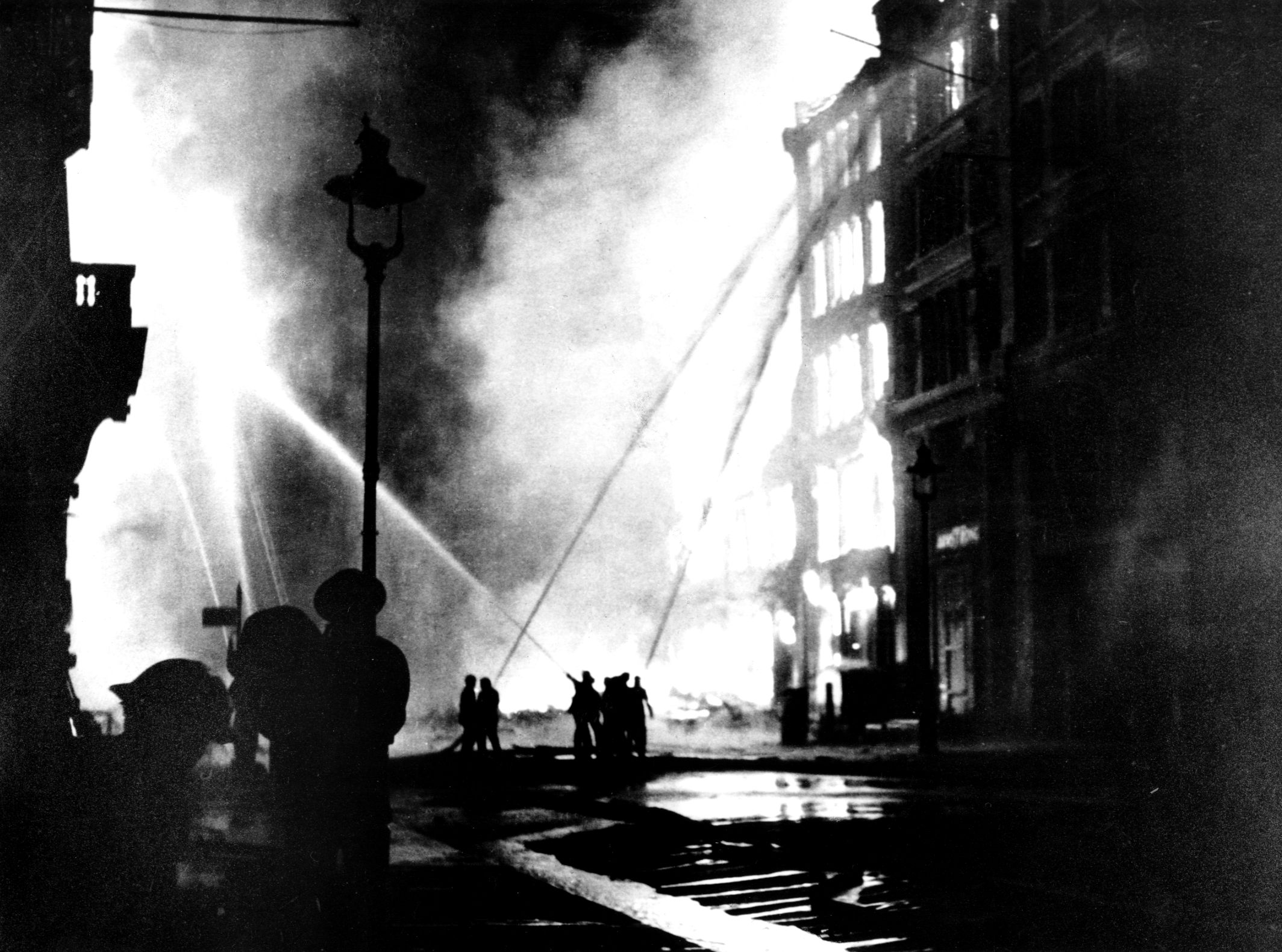 By the end of 1940, nearly 20,000 incendiary bombs had been dropped on London by the Luftwaffe. The London Fire Brigade, several of its members shown here silhouetted against a burning building and directing streams of water toward the flames, performed heroically while battling stricken areas.