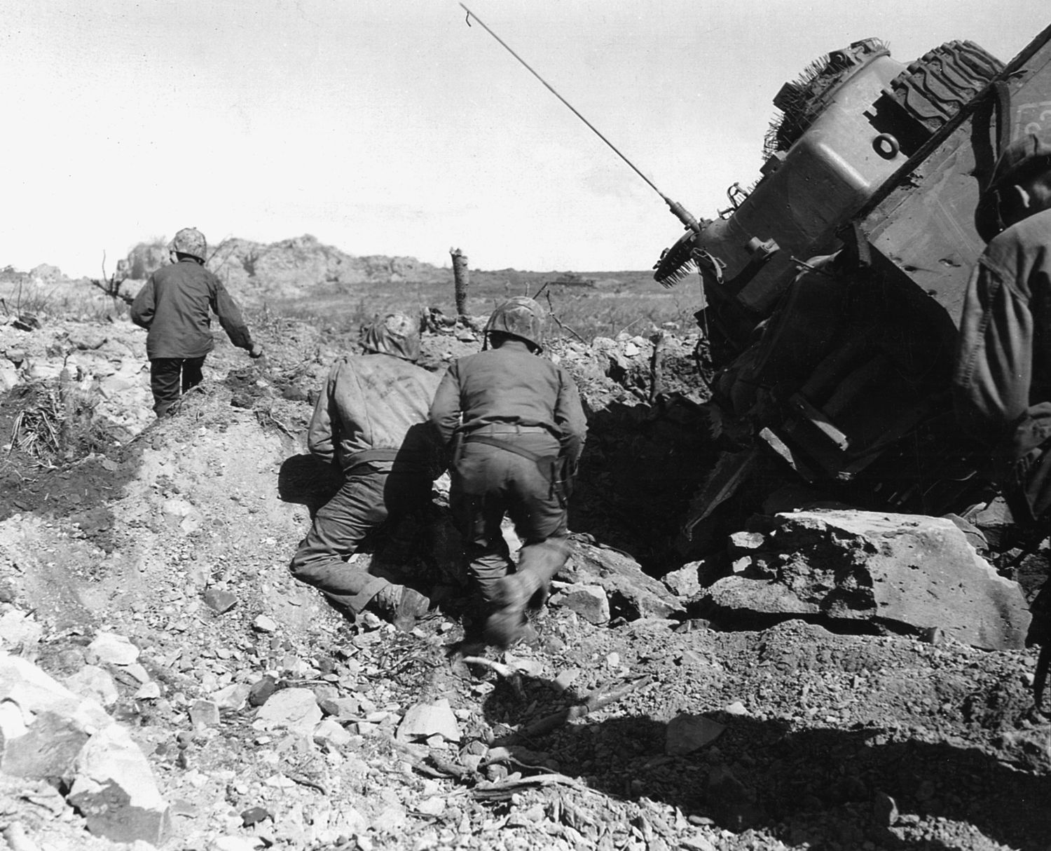 On March 1, 1945, several days after the historic raising of the flag on Iwo Jima, weeks of difficult fighting remained. Here, a squad of U.S. Marines advances past a tank, which has become trapped in the rubble of a demolished Japanese pillbox.