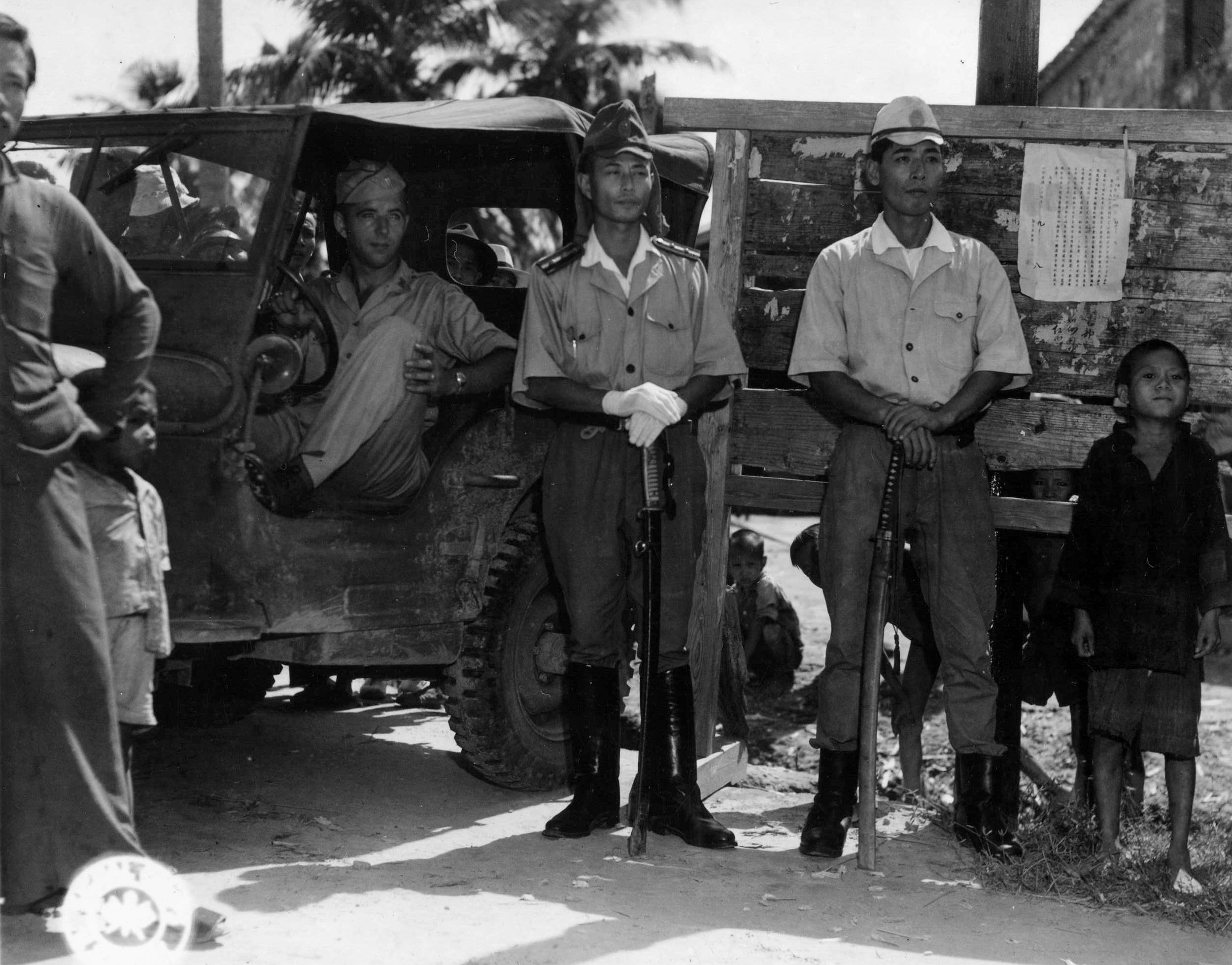 American Lieutenant Frank G. Beiser and two officers belonging to the Japanese Marine contingent on the island of Hainan pose for the camera beside a sign proclaiming the end of the war and urging any escaped Allied prisoners to come in from hiding.