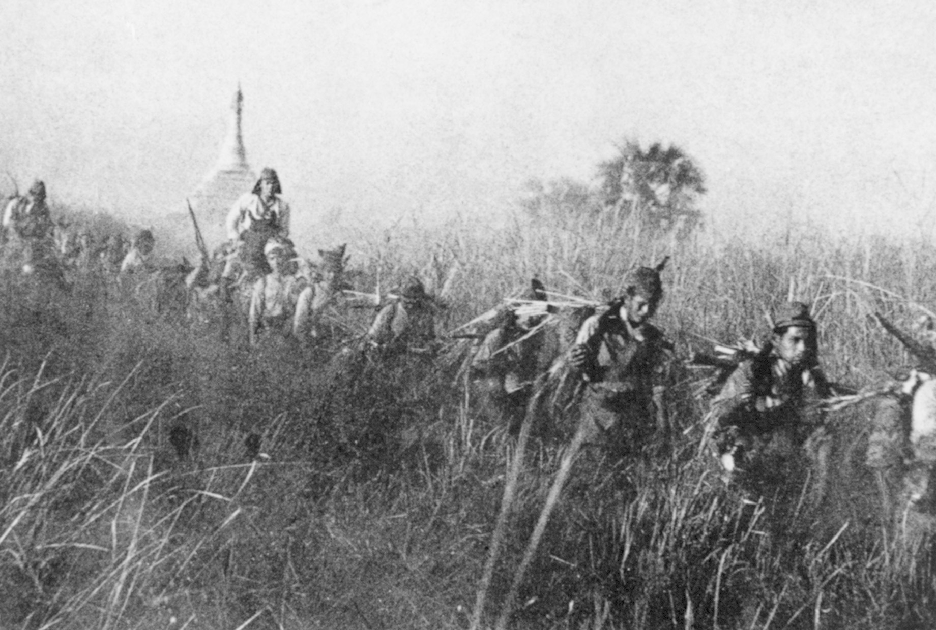Japanese troops make their way through tall grass in the Burmese jungle, some sporting camouflage covering on their backs. Acknowledged as expert jungle fighters, the might of the Japanese Army in Burma was turned back by the tenacious defenders of Kohima. 