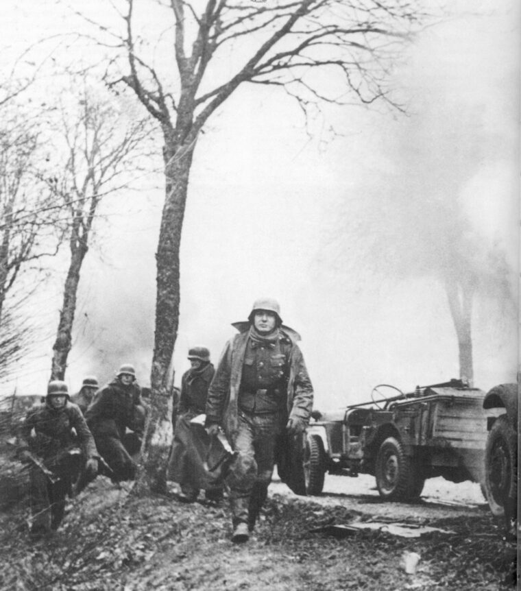 German troops stride past flaming American vehicles during the Battle of the Bulge. The soldier in the foreground carries a Mauser Kar 98 rifle, while the one at left holds a StG44 assault rifle.