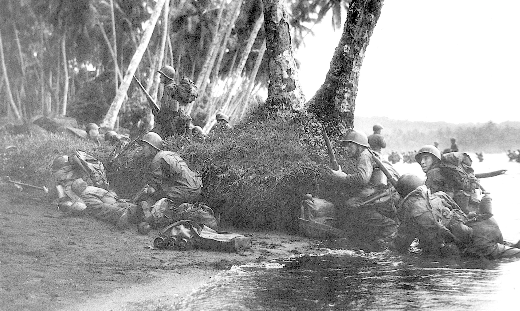 Attacking at the break of day in a heavy rainstorm, the first Marines ashore take cover behind tree trunks and anything else they can find.