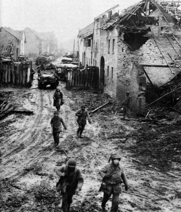 A battleworn column of American troops makes its way through the remains of a village in the Ardennes.
