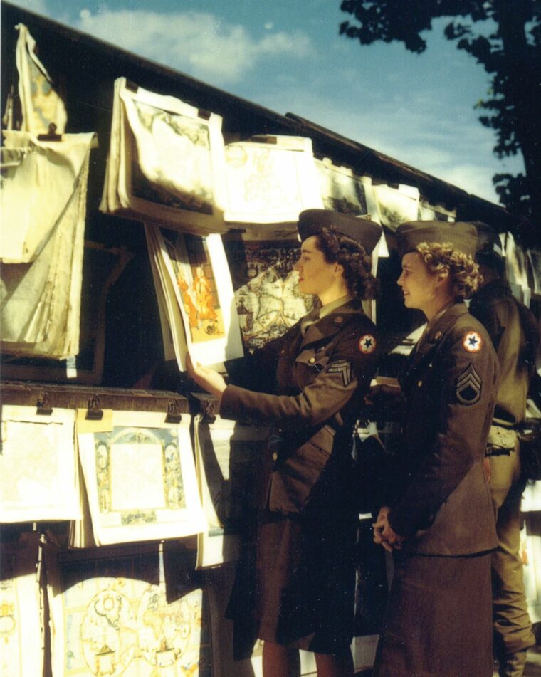 Two women NCOs from the Army’s supply service headquarters examine some renaissance lithographs from an open-air art peddler.