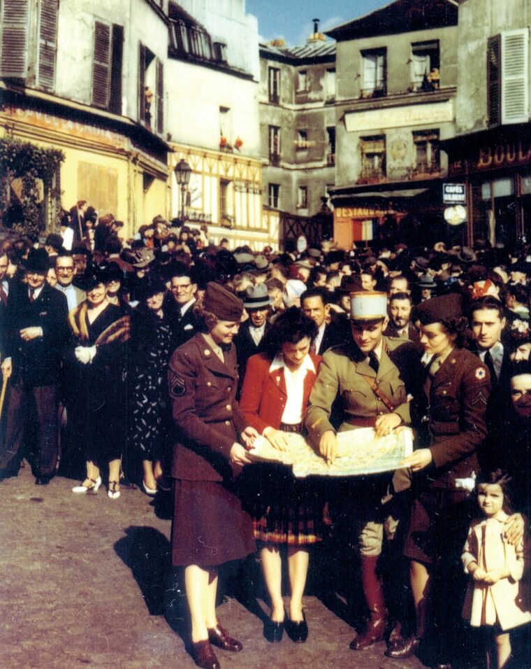 On a crowded street, a French Arm officer and a French woman help two American NCOs find themselves on a map.