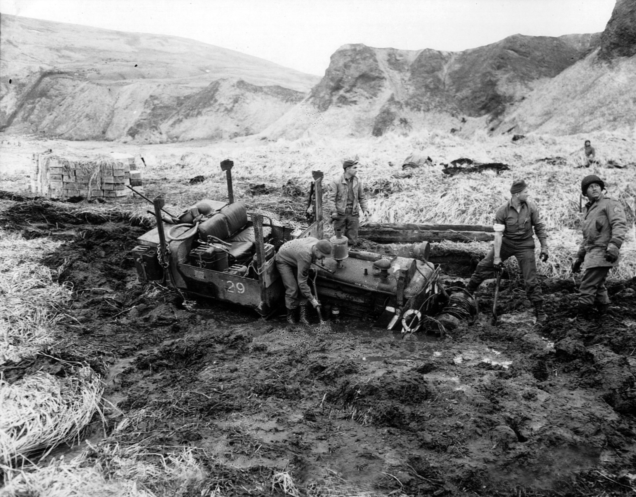Bogged down on a muddy trail in the Aleutians, a tractor meant to move earth has become its prisoner. The sound of approaching aircraft has grabbed the attention of the soldiers attempting to reclaim the equipment.