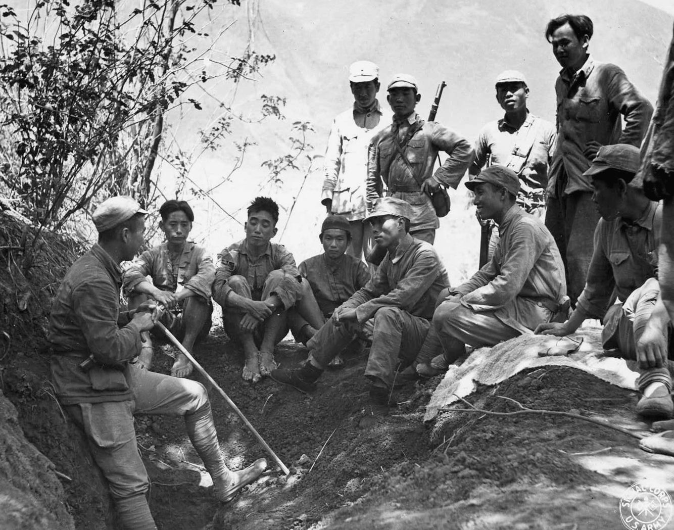 Major Chu, a Chinese officer, confers with a group of soldiers and individuals who have volunteered to gather intelligence for use in upcoming operations. Chu is instructing the men from the relative safety of a dugout 200 feet from the banks of the Salween River.