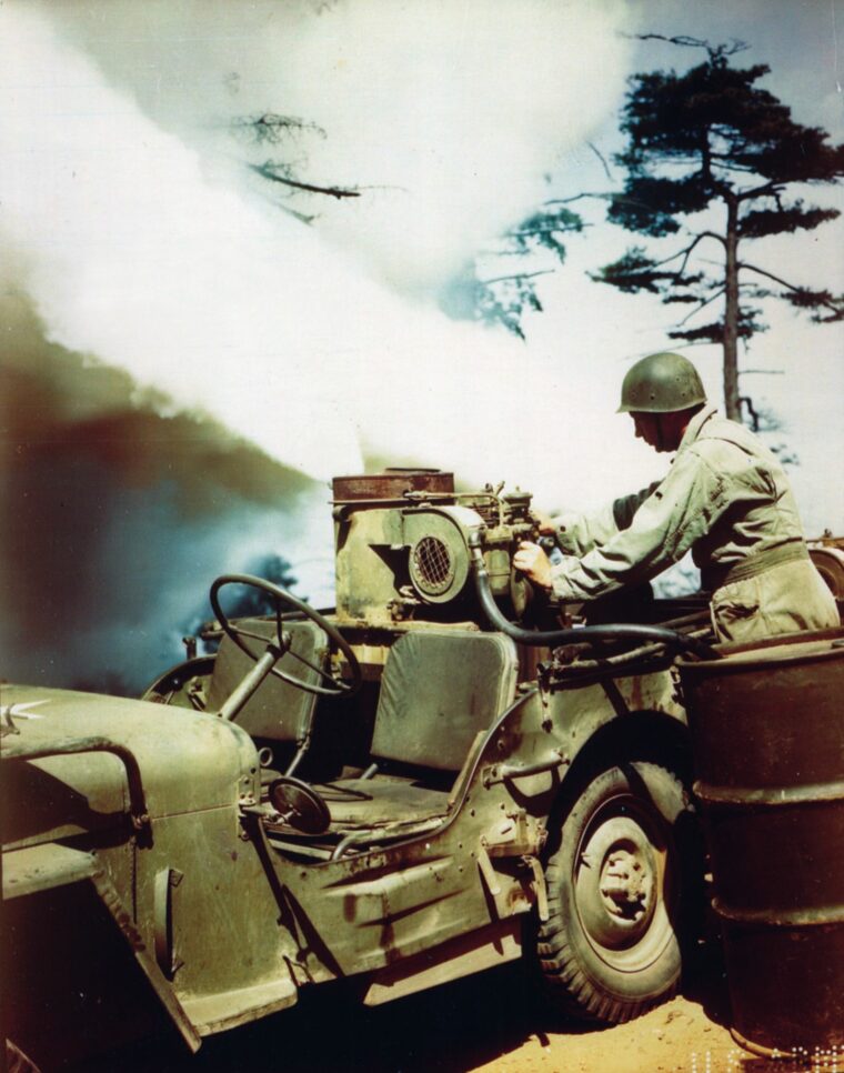 Screening an advancing Army column, an M-2 smoke generator mounted on a Jeep belches a plume of chemical fog.