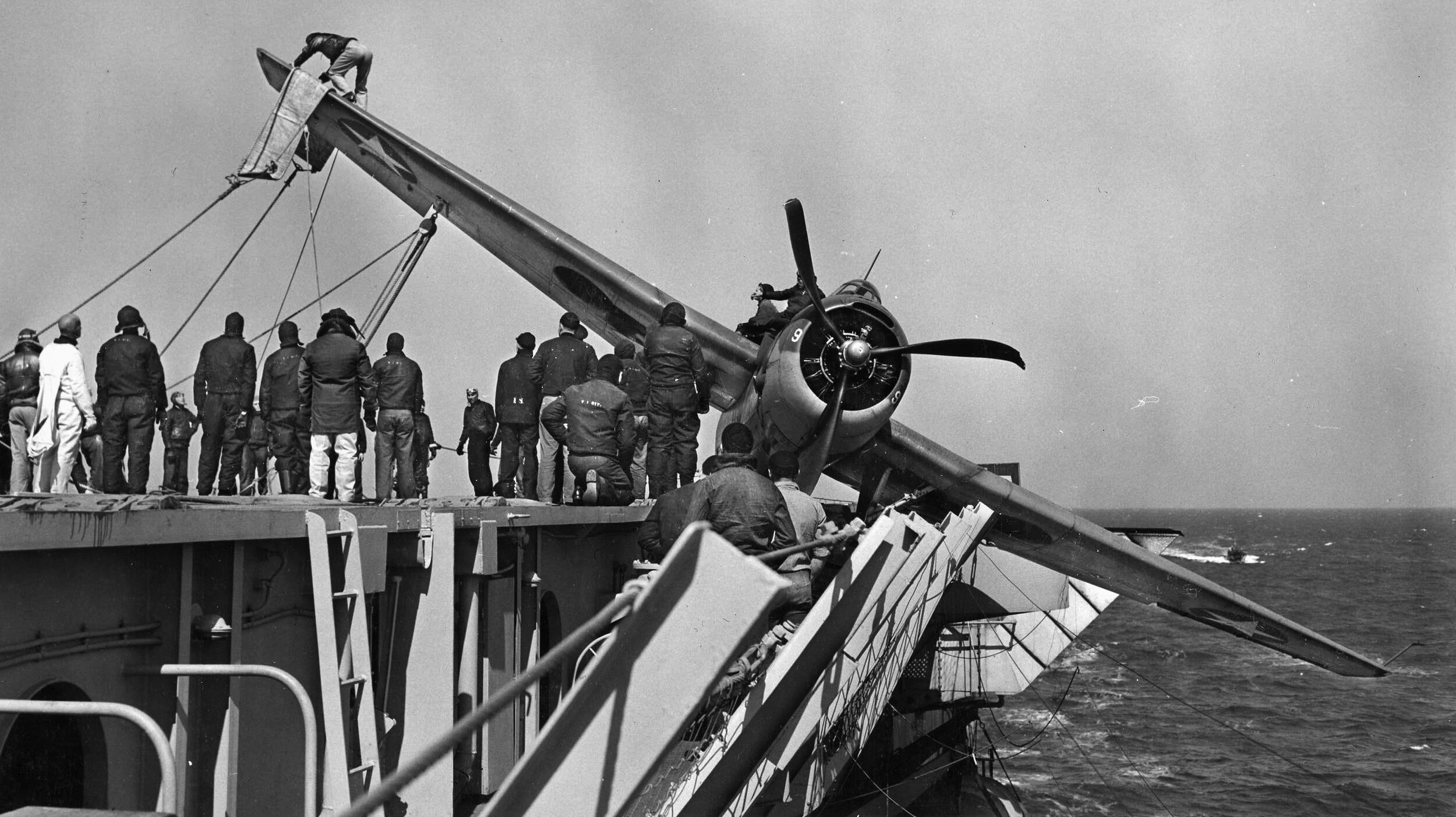 Using winches and a sling, Navy deck hands upright a TBF Avenger torpedo bomber that crashed on landing. The bent propeller attests to a harsh landing.