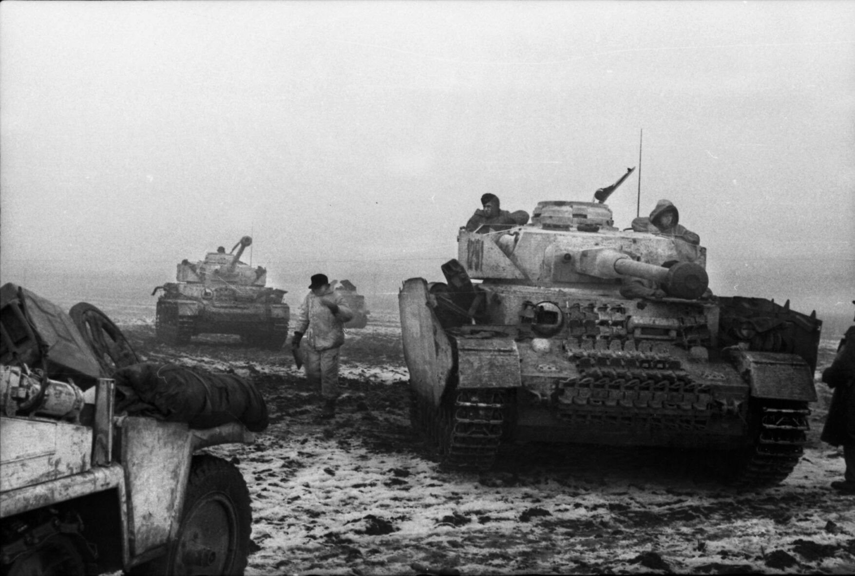 Two German PzKpfw. IV tanks advance along a bleak, muddy landscape in December 1943, as panzergrenadiers trudge down a Ukrainian road that has turned into a quagmire or hitch rides on the tanks. Note the side armor on the vehicles, intended to detonate antitank shells prior to impact with the tank’s hull.