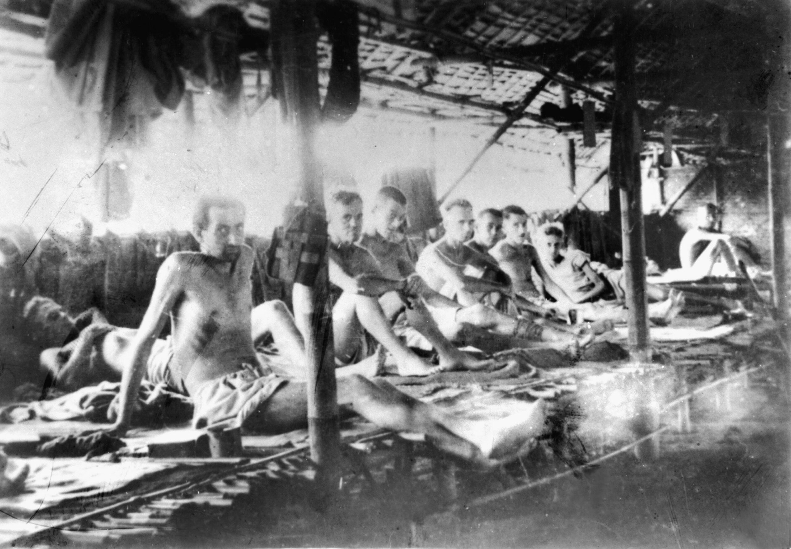 Emaciated Allied prisoners stare blankly at the photographer who snapped this photo of them lying on sleeping platforms in a large POW camp hut.