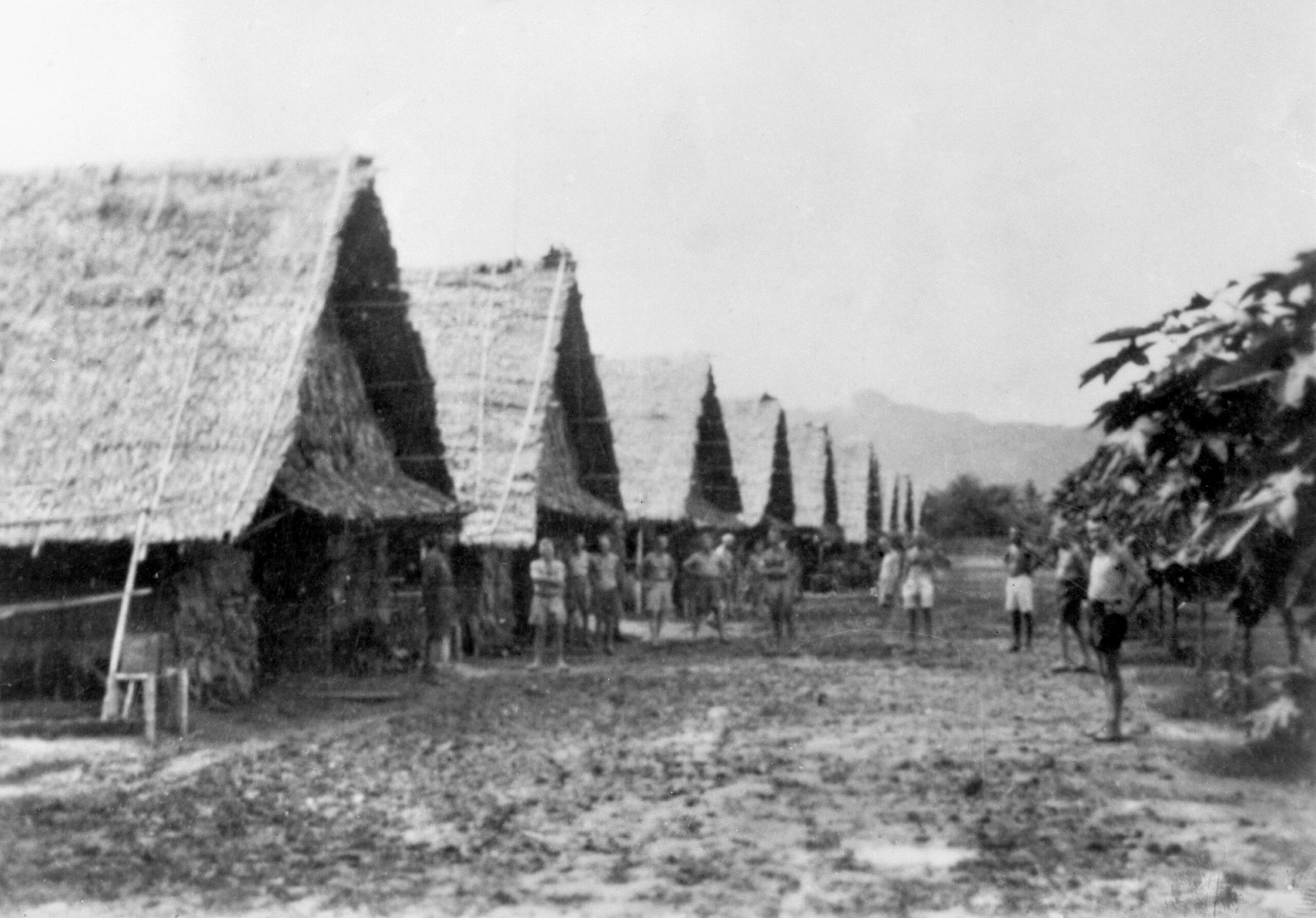 Allied prisoners stand outside their huts at the Nong Pladuk POW camp. Charlie Mott was sent to this location in September 1942. After the completion of the Burma-Thailand Railway, a number of prisoners were taken to Nong Pladuk. Others boarded Hell Ships for the arduous trek to Japan. The headquarters building of the Japanese camp commander can be seen in the background.