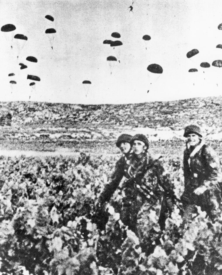 Glancing toward their photographer, three German paratroopers, wearing their distinctive rimless helmets, march off toward an assembly area while other fallschirmjager land in the field beyond. The capture of the airfields finally gave the invaders the upper hand on Crete.