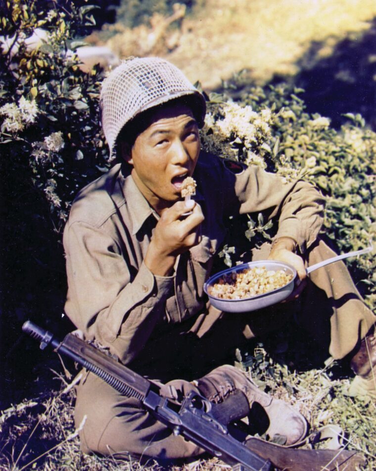 A Japanese American soldier with the 100th Infantry Battalion rests his Thompson machine gun so he can enjoy some hot food in Italy. 