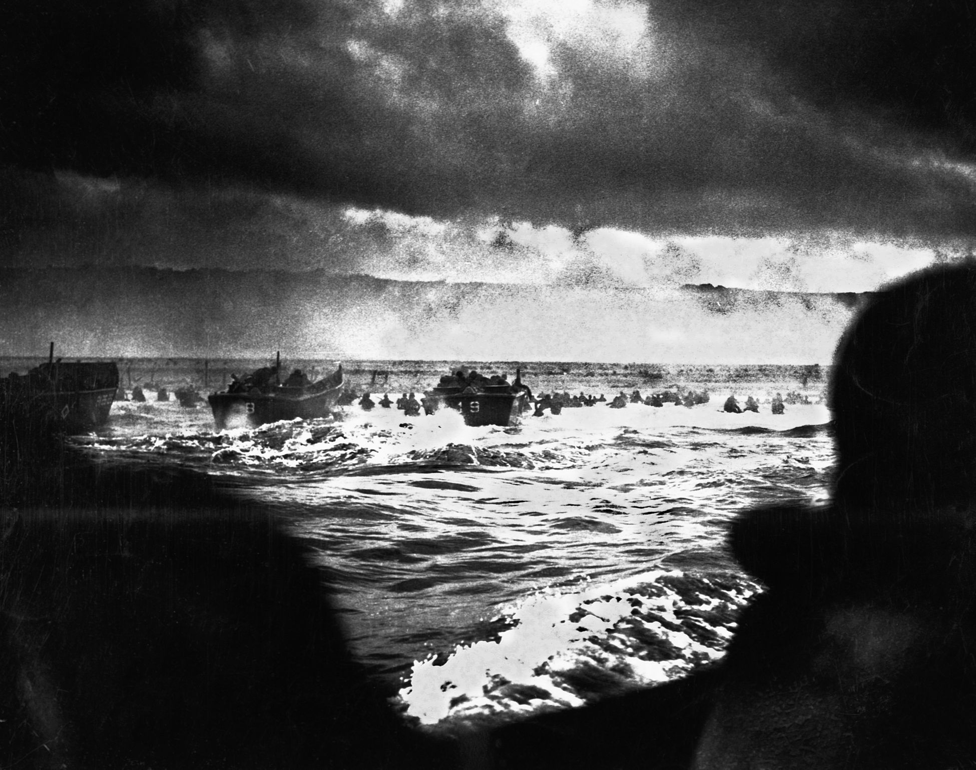 Churning through the rough waters of the English Channel, Higgins boats laden with U.S. soldiers approach Omaha Beach on D-Day. Some soldiers have already disembarked and are seen wading through the surf toward shore, while intense German fire rakes the waterline. Shrouds of smoke are visible in the distance.