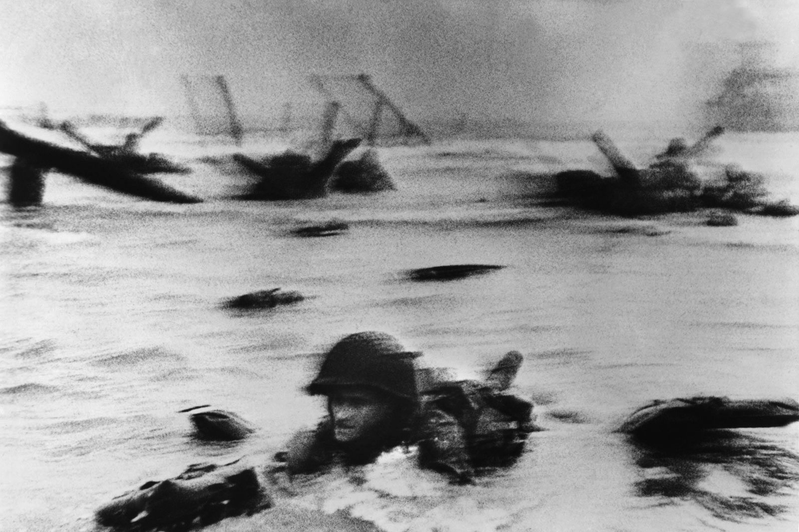 In the above photo, a soldier struggles through the churning surf toward cover. In the photo on the opposite page, soldiers utilize heavy beach obstacles as refuge against withering German fire directed from the heights above Omaha Beach.