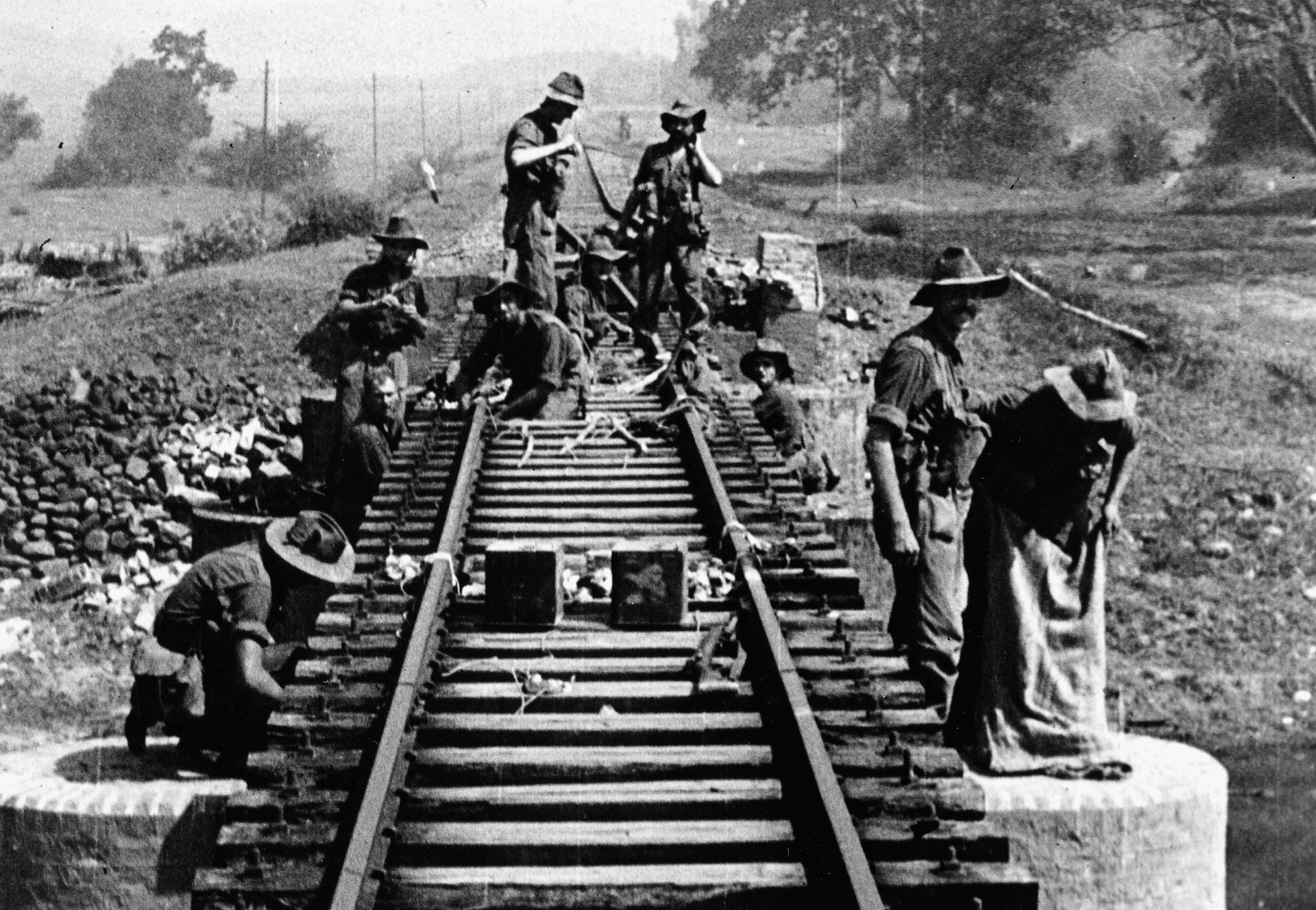 Wearing large-brimmed hats to ward off the tropical sun, engineers of the 77 Brigade wire a section of railroad track with explosives.