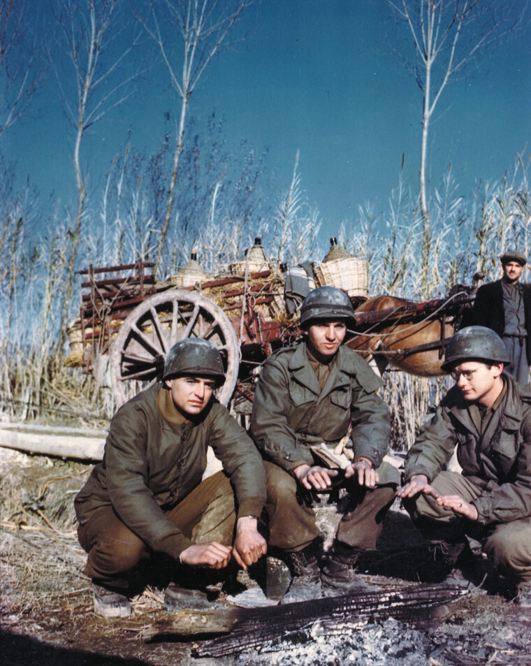 Staving off the bitter cold, three U.S. soldiers of the 85th Infantry Division huddle around a fire as a curious Italian villager looks on.