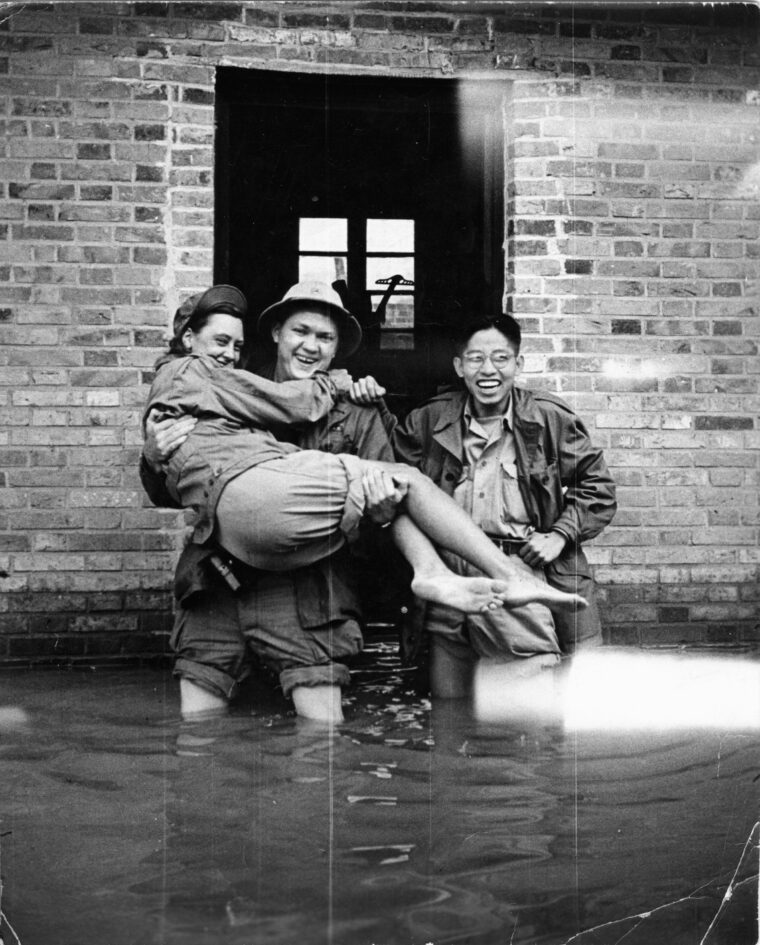Amid heavy flooding in 1945, Elizabeth McIntosh is carried to high ground by Sergeant William A. Smith. Famout Chinese cartoonist Tong Ting smiles broadly as the trio exits the Morale Operations print shop.