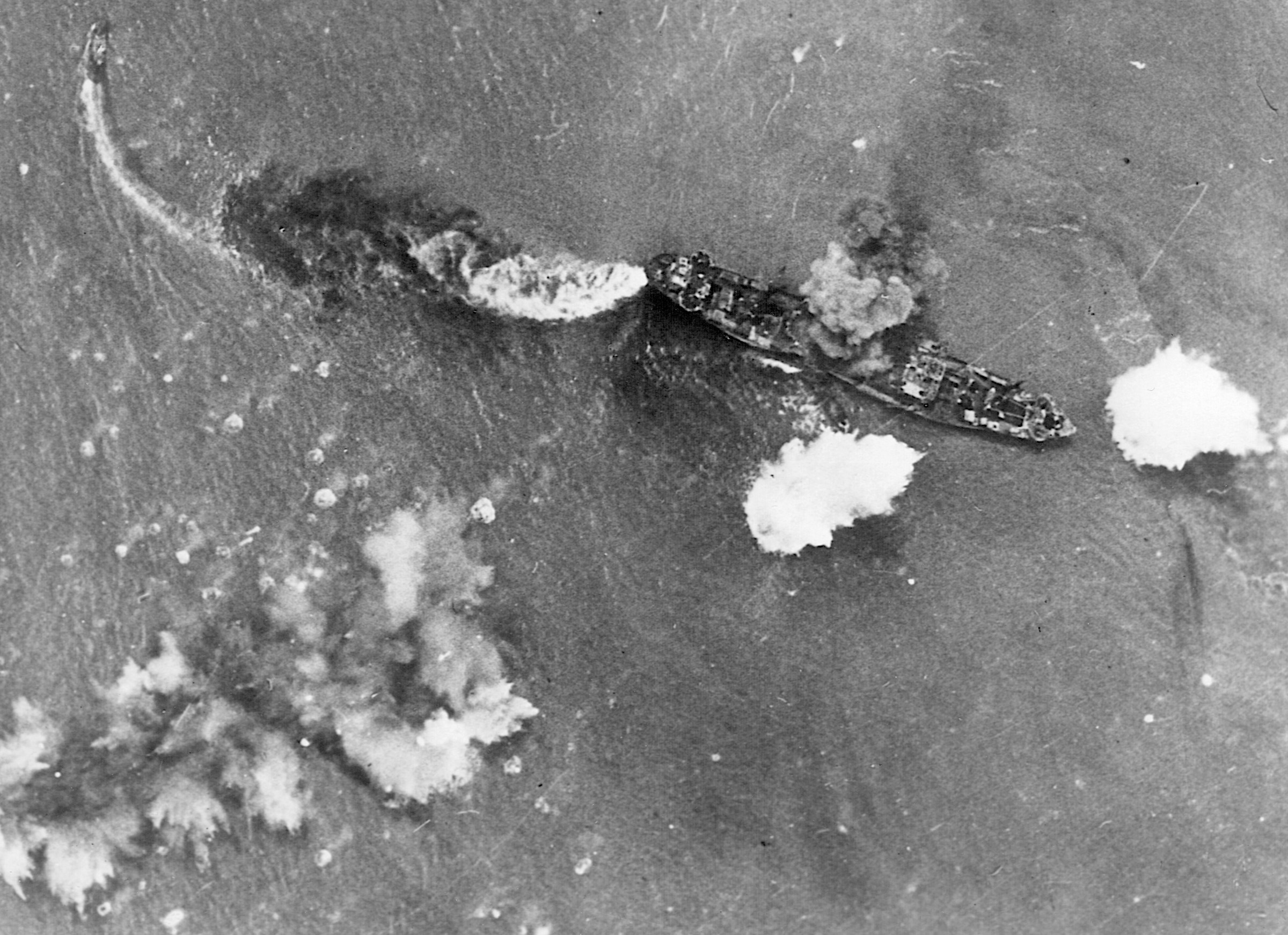A stricken Japanese vessel billows smoke from a bomb hit amidships while a fire rages in the stern. Moments later, the transport settled to the bottom of the Bismarck Sea. A smaller boat attempts to escape the debacle.