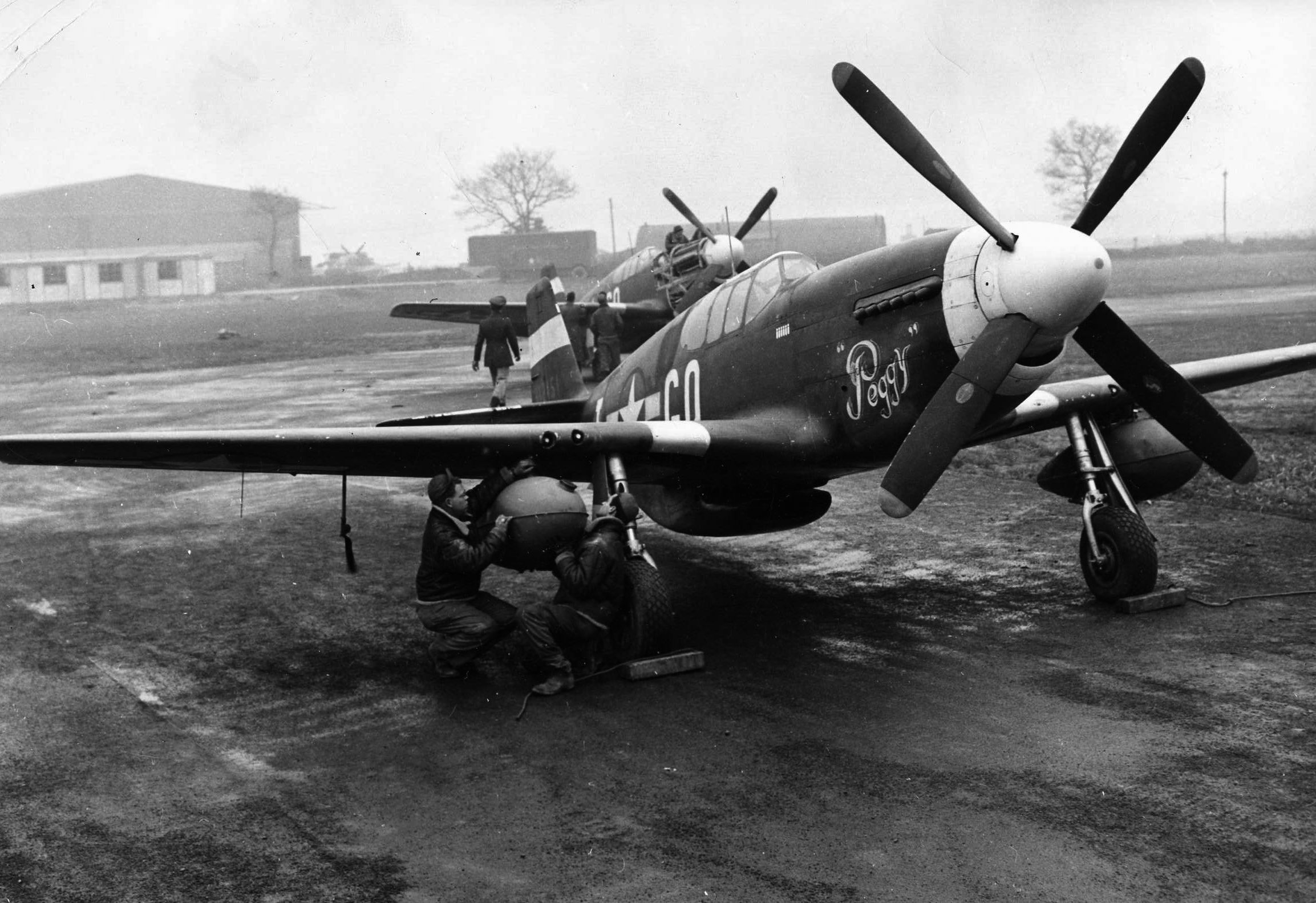 On a muddy airfield in England, P-51 Mustangs sit ready for their pilots to climb aboard as two ground crewmen make a final check of an external drop tank.  The extra range provided by the drop tanks allowed the fighters to escort bomber formations deeper into Germany.